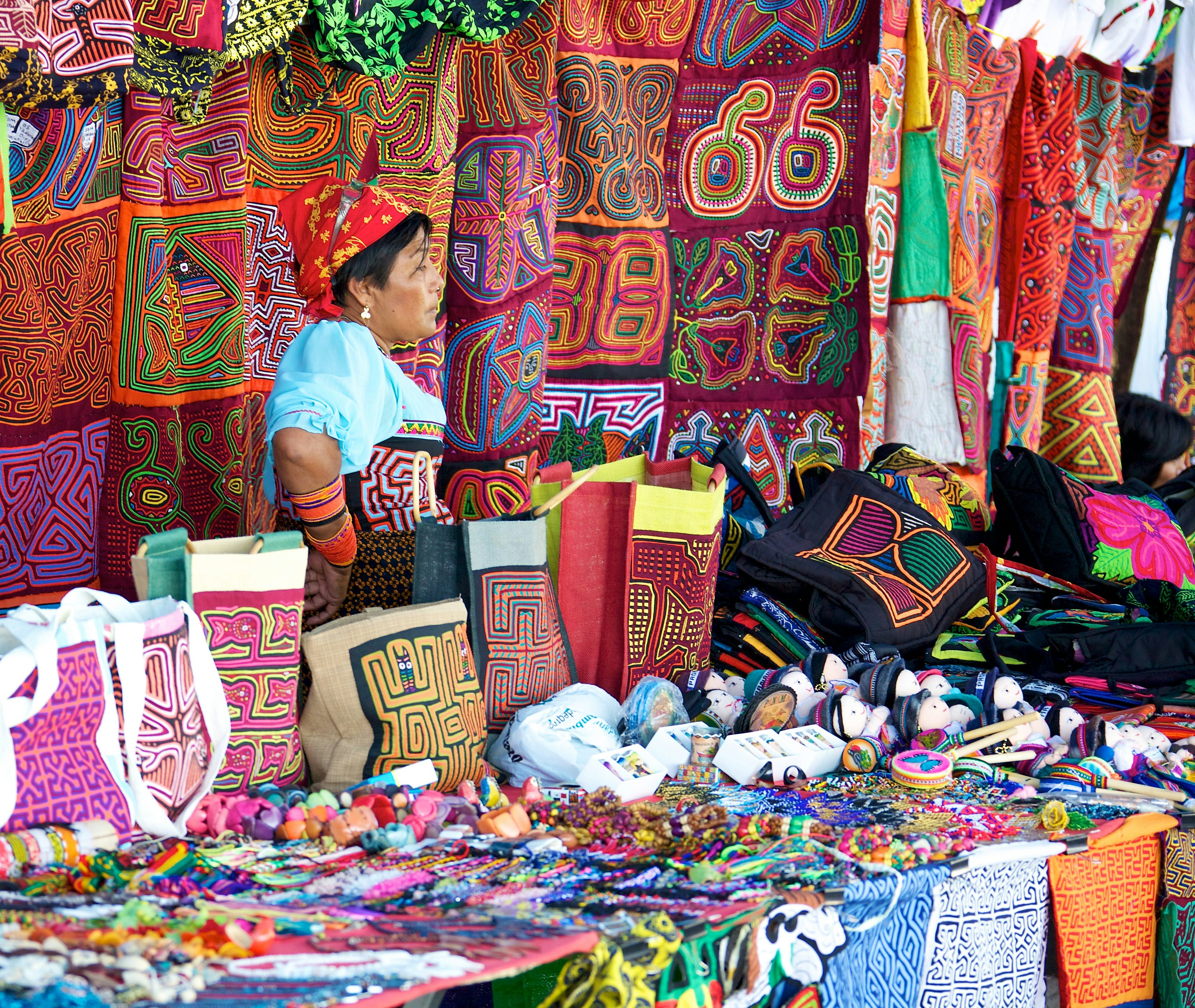 Woman standing behind a table of items made from colorful fabrics as well as colorful jewelry. More colorful fabrics hang behind her.