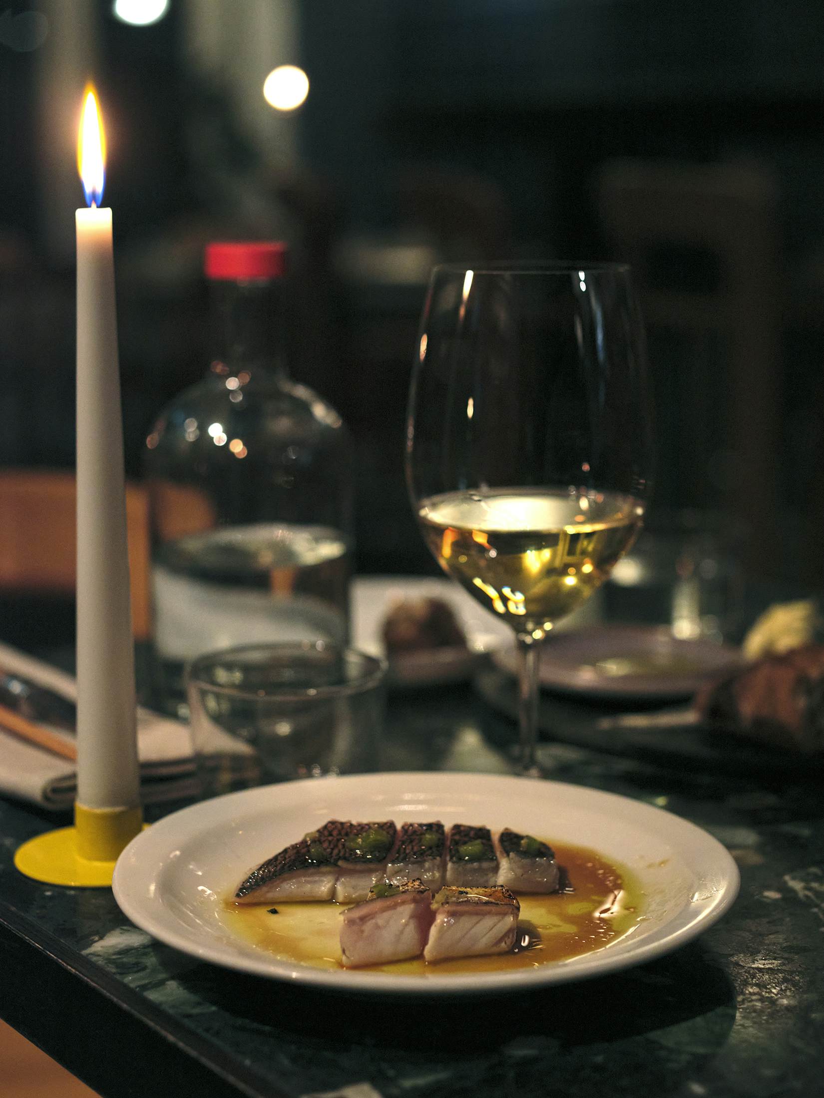 A glass of white wine and a small plate with fish dressed in olive oil are on a candlelit table.