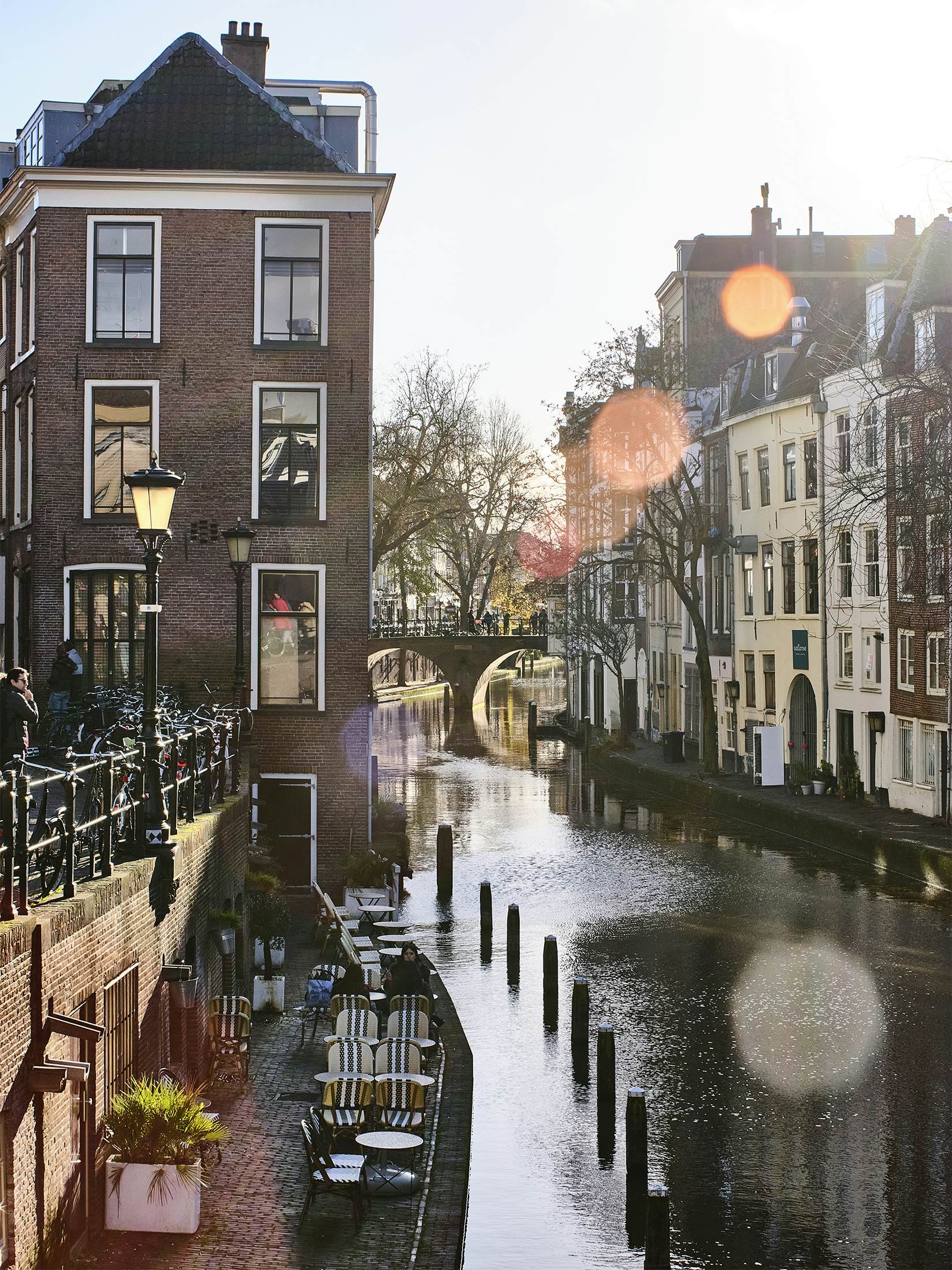 A split-level canal. On ground level, bicycles are parked at a railing; at the water level, empty outdoor tables sit beside the water.