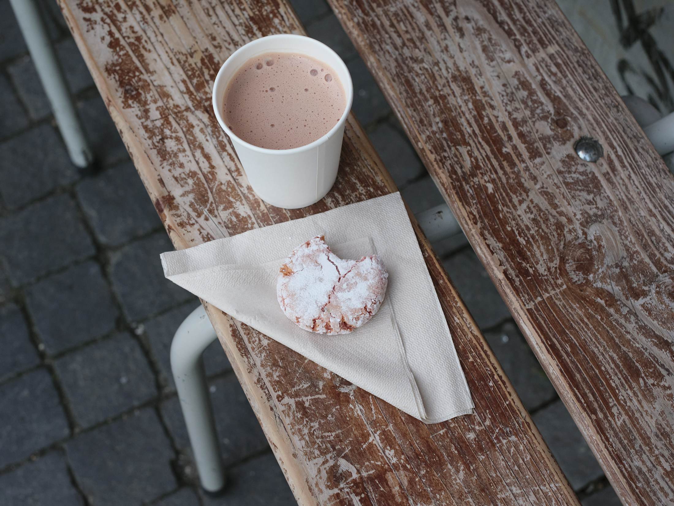 Hot chocolate in a paper cup and a cookie on a folded paper napkin rest on a bench.
