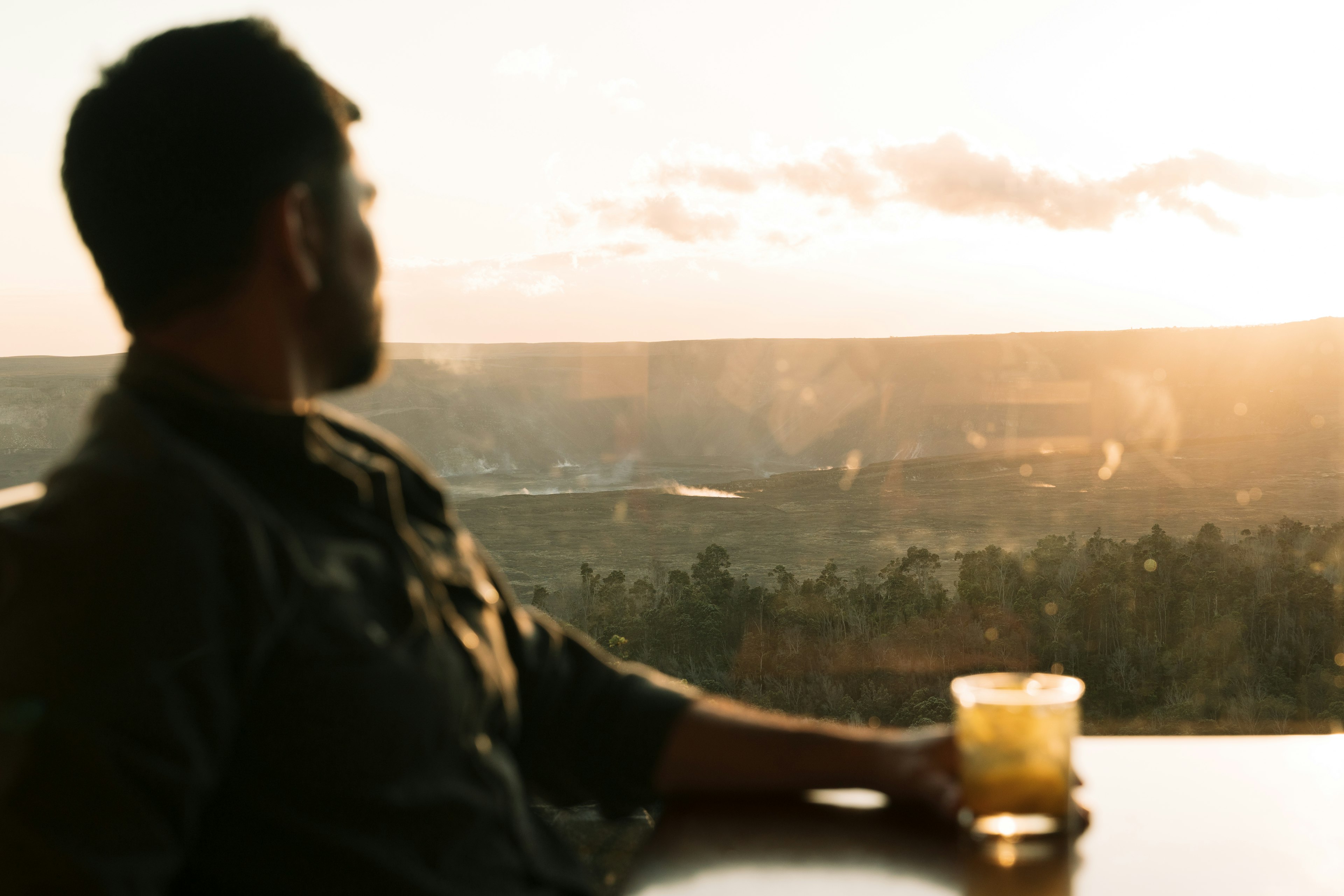A man looks out a window at sunset while he sits at a table with a cocktail