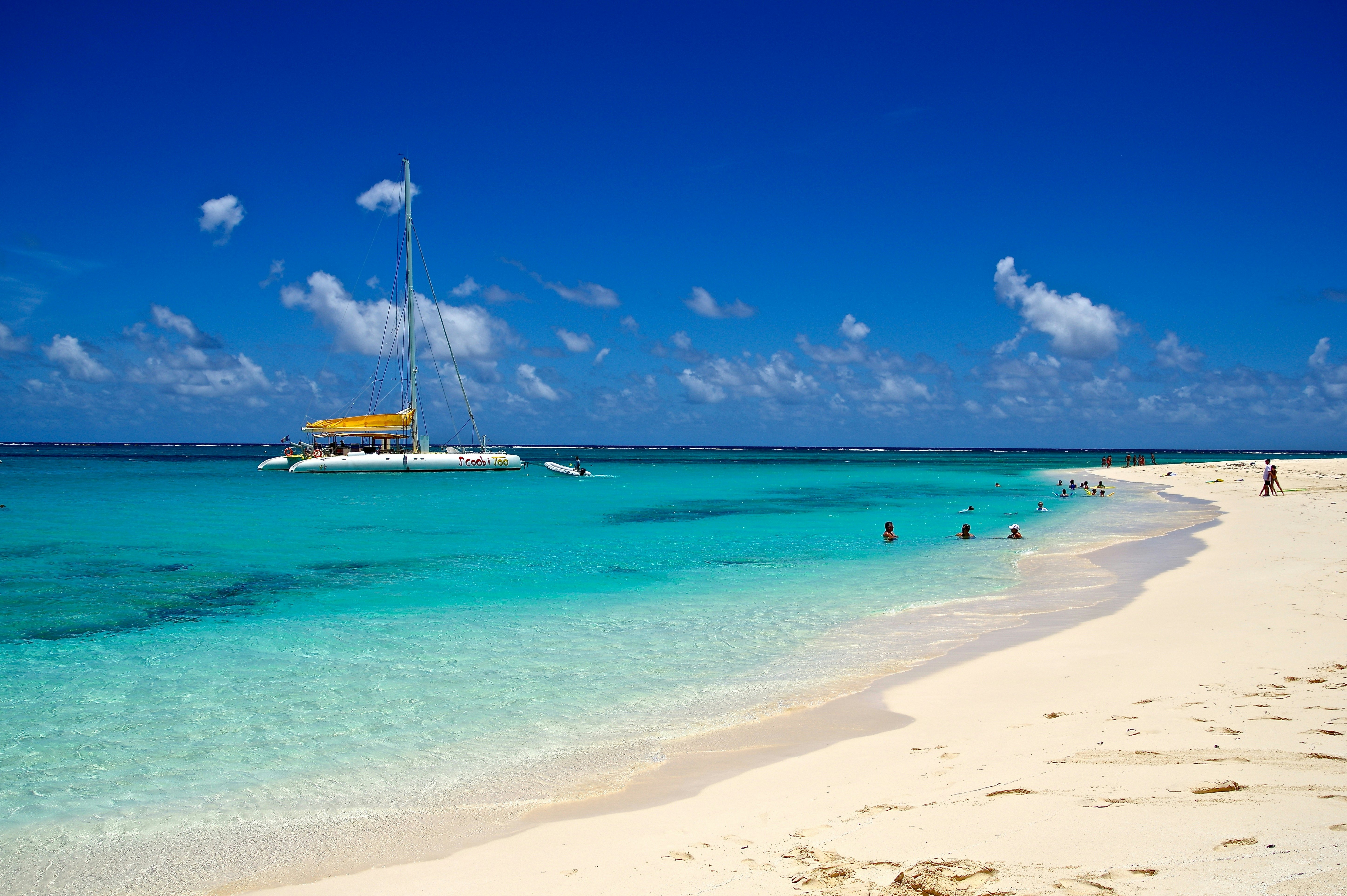 A catamaran moored in turquoise waters off the shore of a sandy beach. Some people lounge in the shallows.
