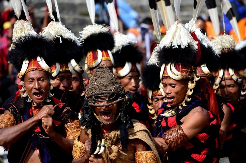 A group of men wearing the traditional clothes of their tribe and a black and white headdress with a white feather at the top stand together as a group at a festival.