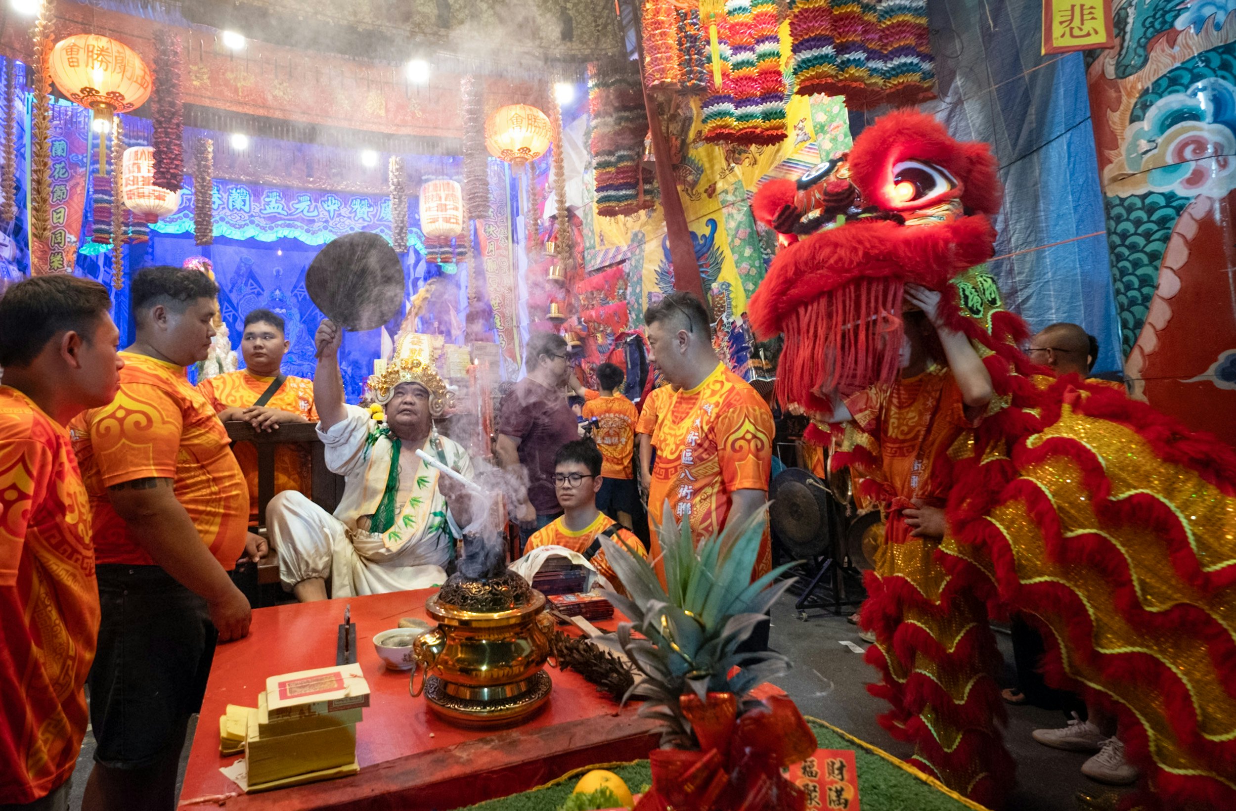 People in orange shirts surround a smoking gold urn while a man in white with a gold crown fans the smoke a chinese dragon puppet is to the right.