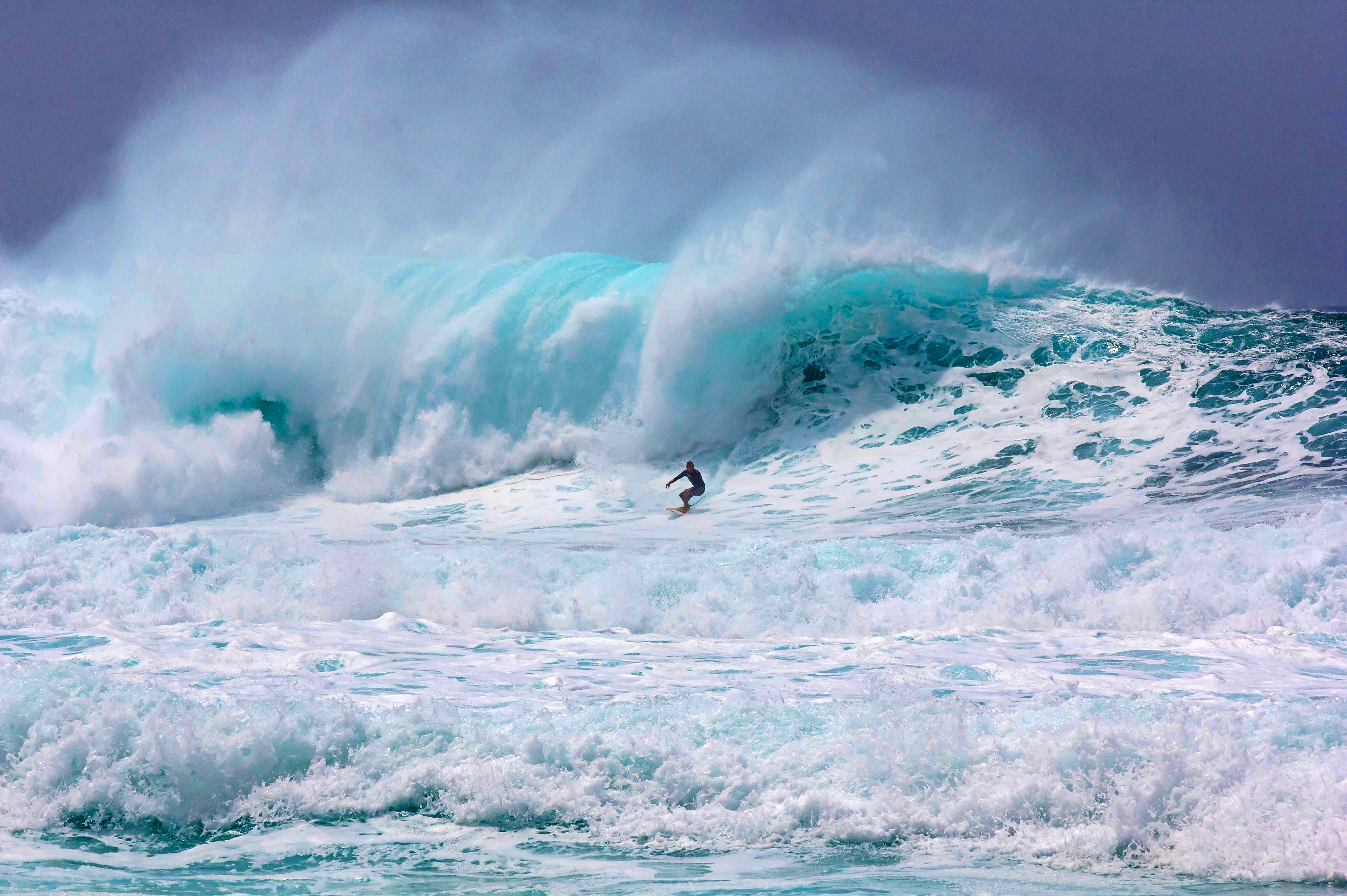 A single surfer rides through white foamy water with a wave crashing behind them.