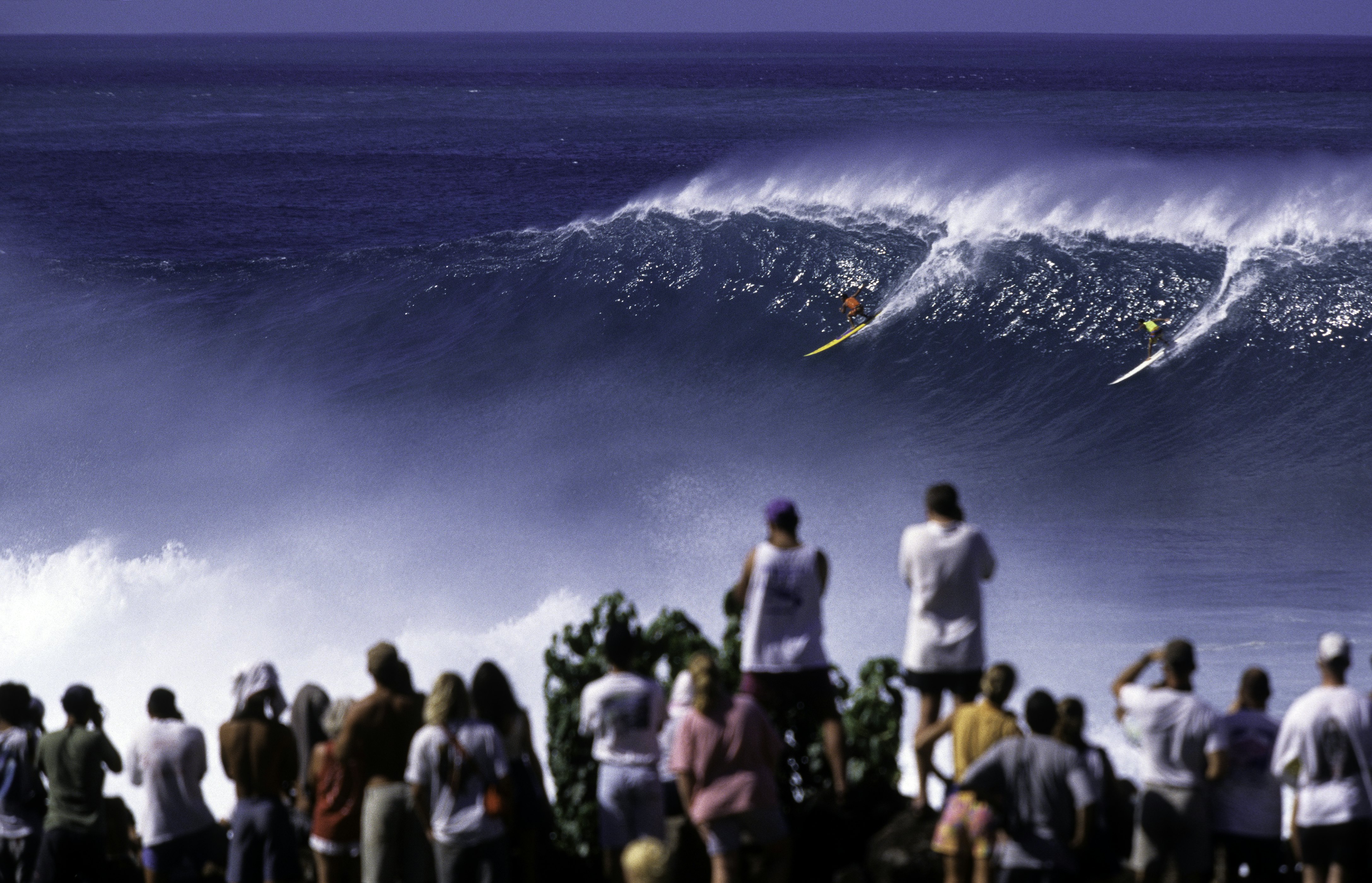 A crowd of onlookers in the foreground watch two surfers at the very top of a giant wave.