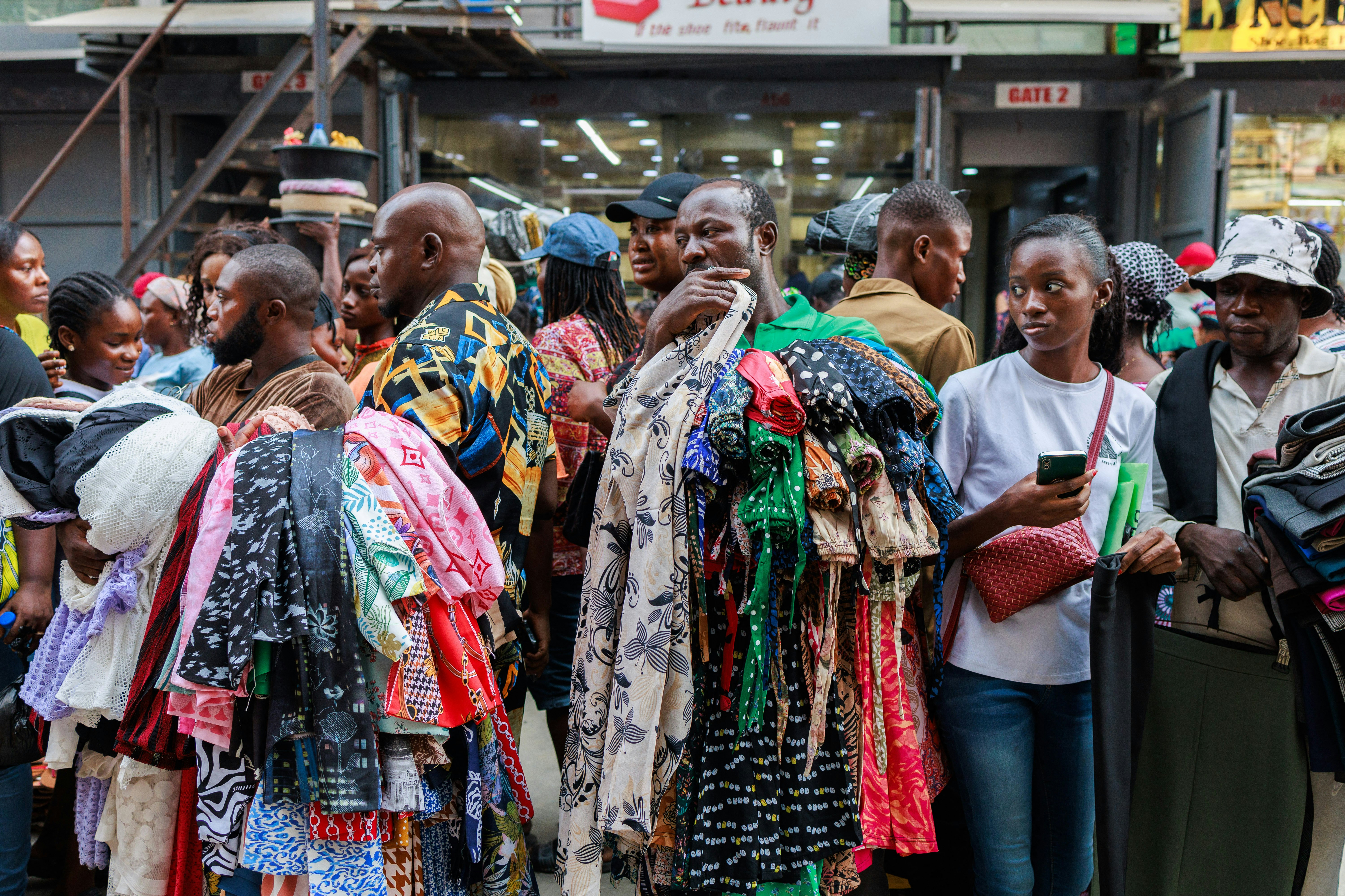 Two men hold piles of printed textiles in a crowded street market. A young woman looks at them with skepticism.