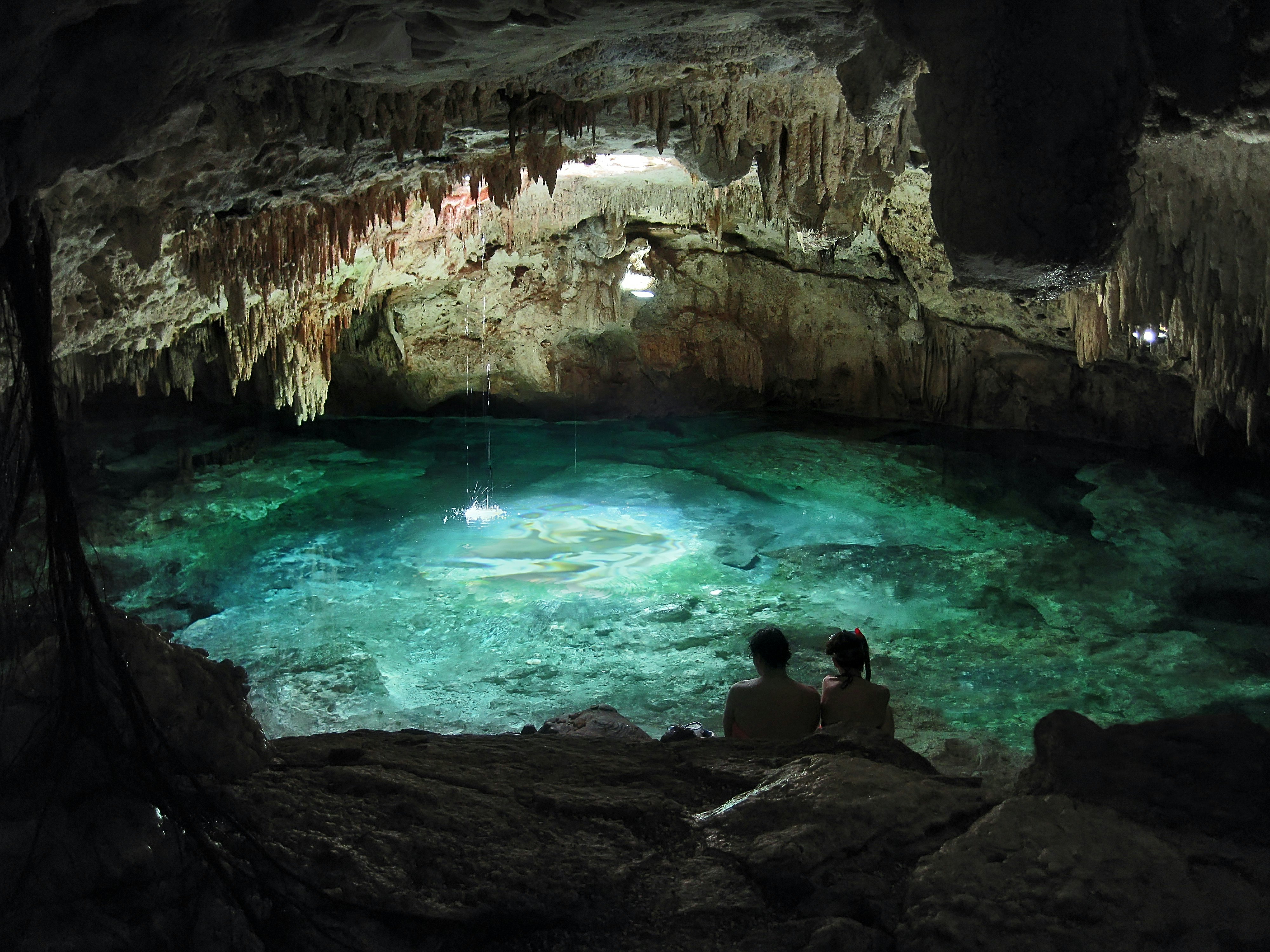 Couple sitting on the rocks (seen from the back) alongside a turquoise underground pond in an open cave with stalagtite concretions on the ceiling.