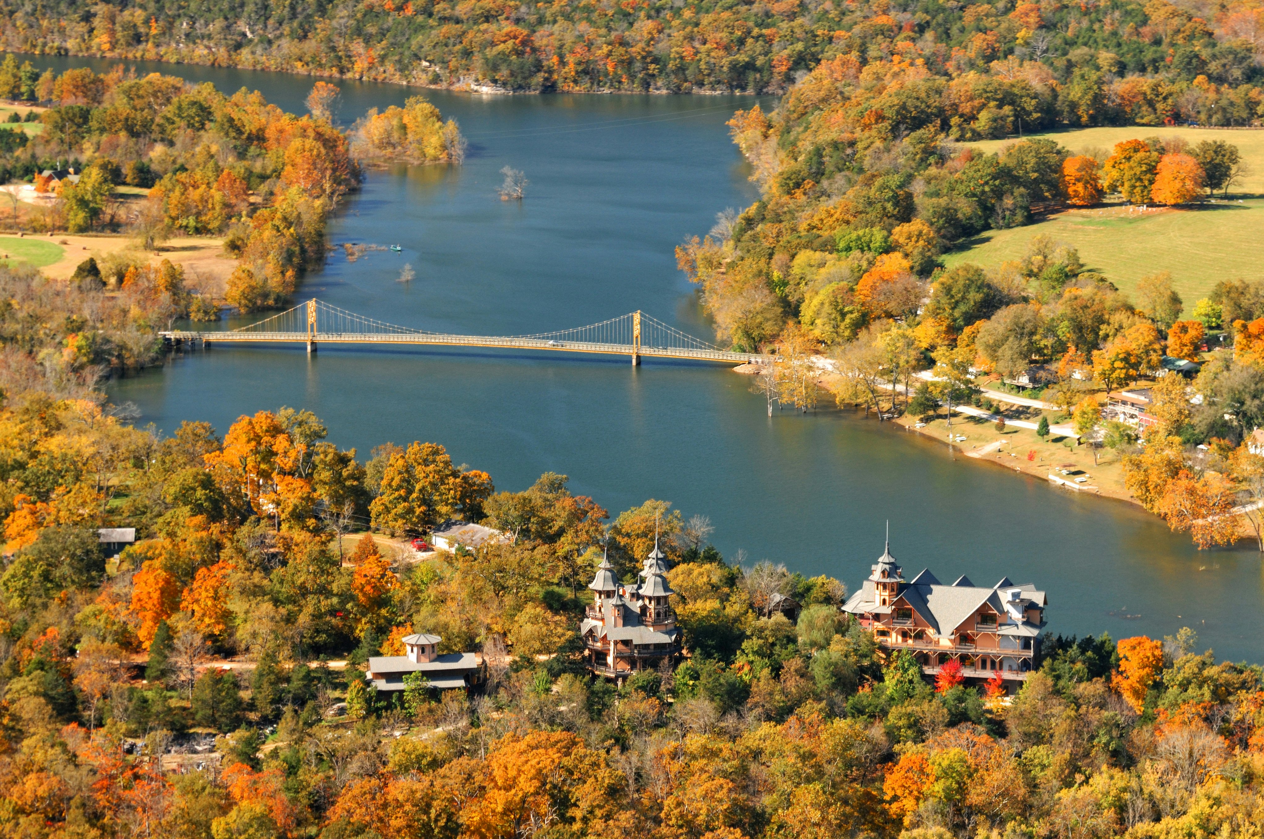 A small riverside settlement with woodland in golden fall colors. Cars drive over a suspension bridge that crosses the wide river.