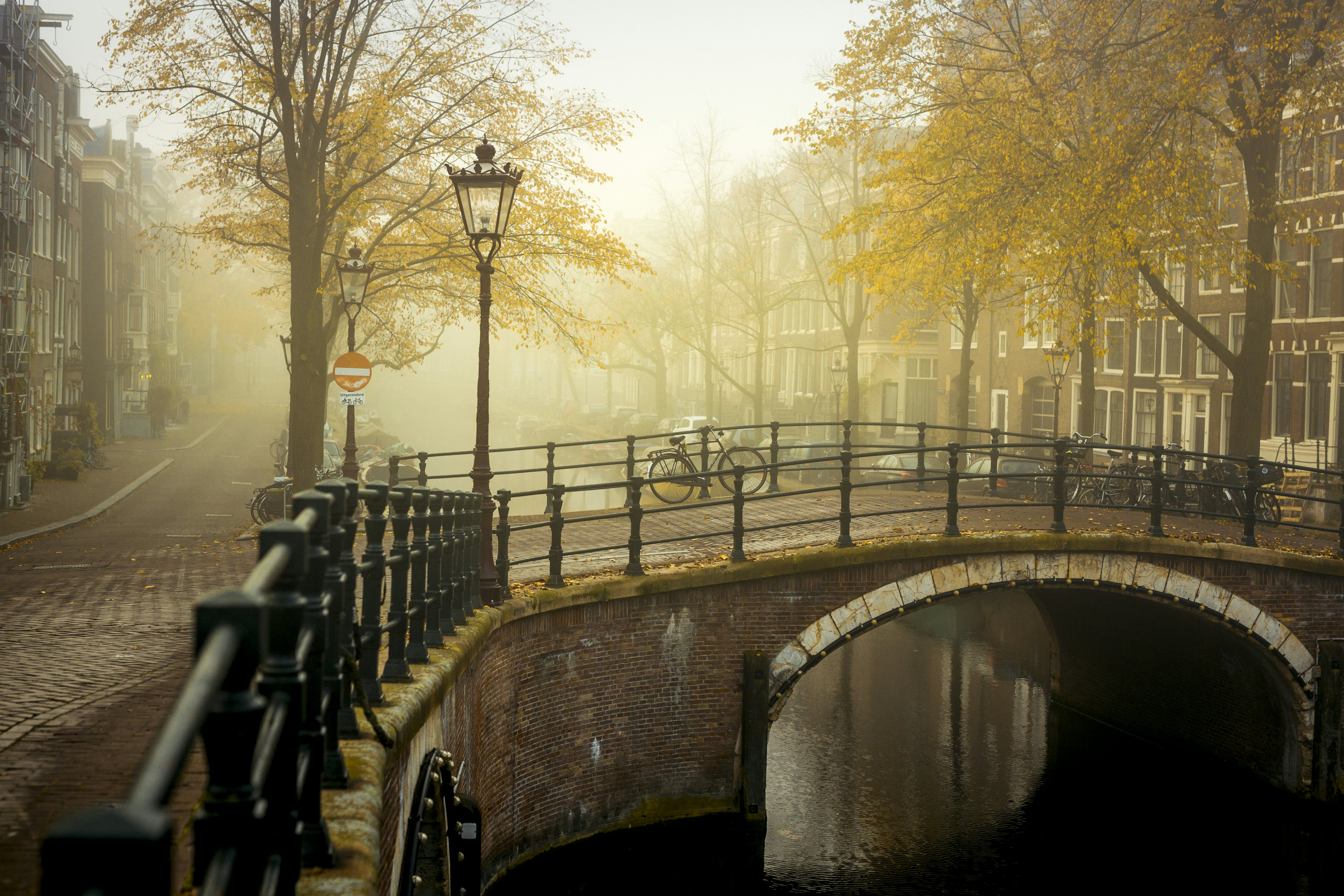 A brick city curve bridge over a city canal on a foggy morning with a lamp post and bicycle. There are brick rowhouses on the streets on either side of the bridge.