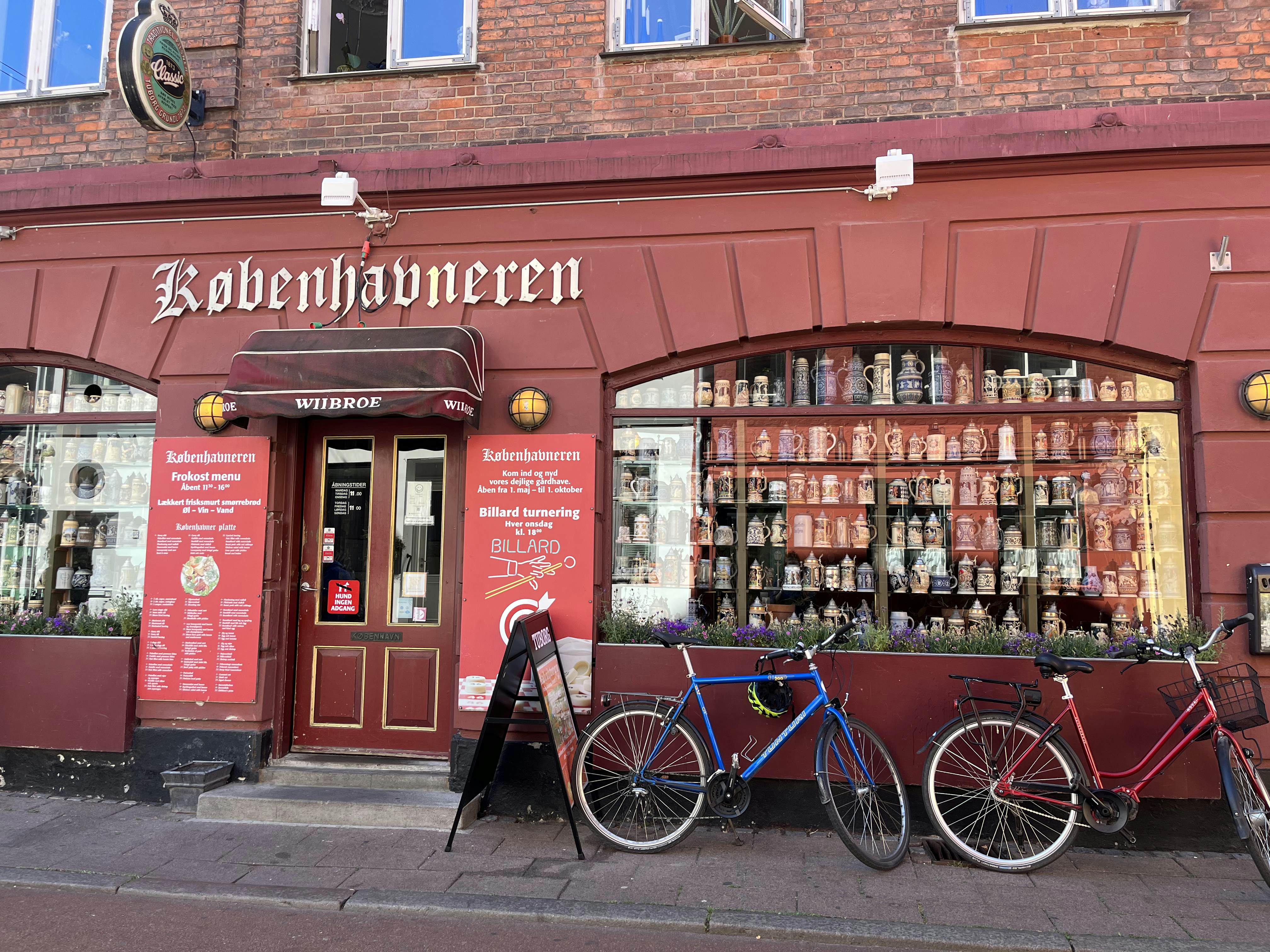 A red storefront with steins in the window and bicycles parked out front on a sunny day.