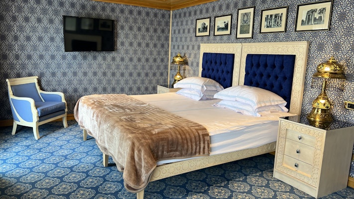 Blue pattered wallpaper covers the walls of a room equipped with a large bed with a blue cushioned headboard. Two gold plated lamps are next to it, and the carpet is also a blue pattern