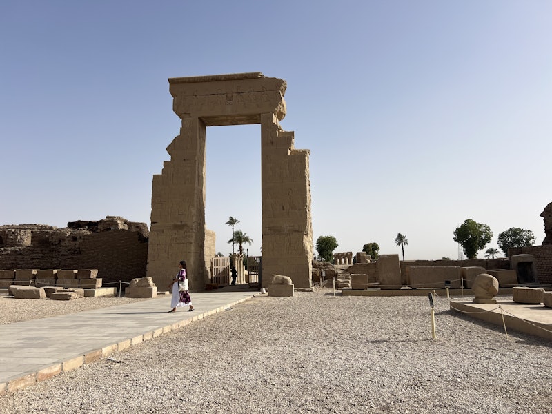 A stone archway decorated with ancient art through which you can see two palm trees. A woman walks in front