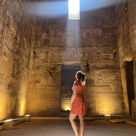 A woman in a stone room looks through a square hole of light