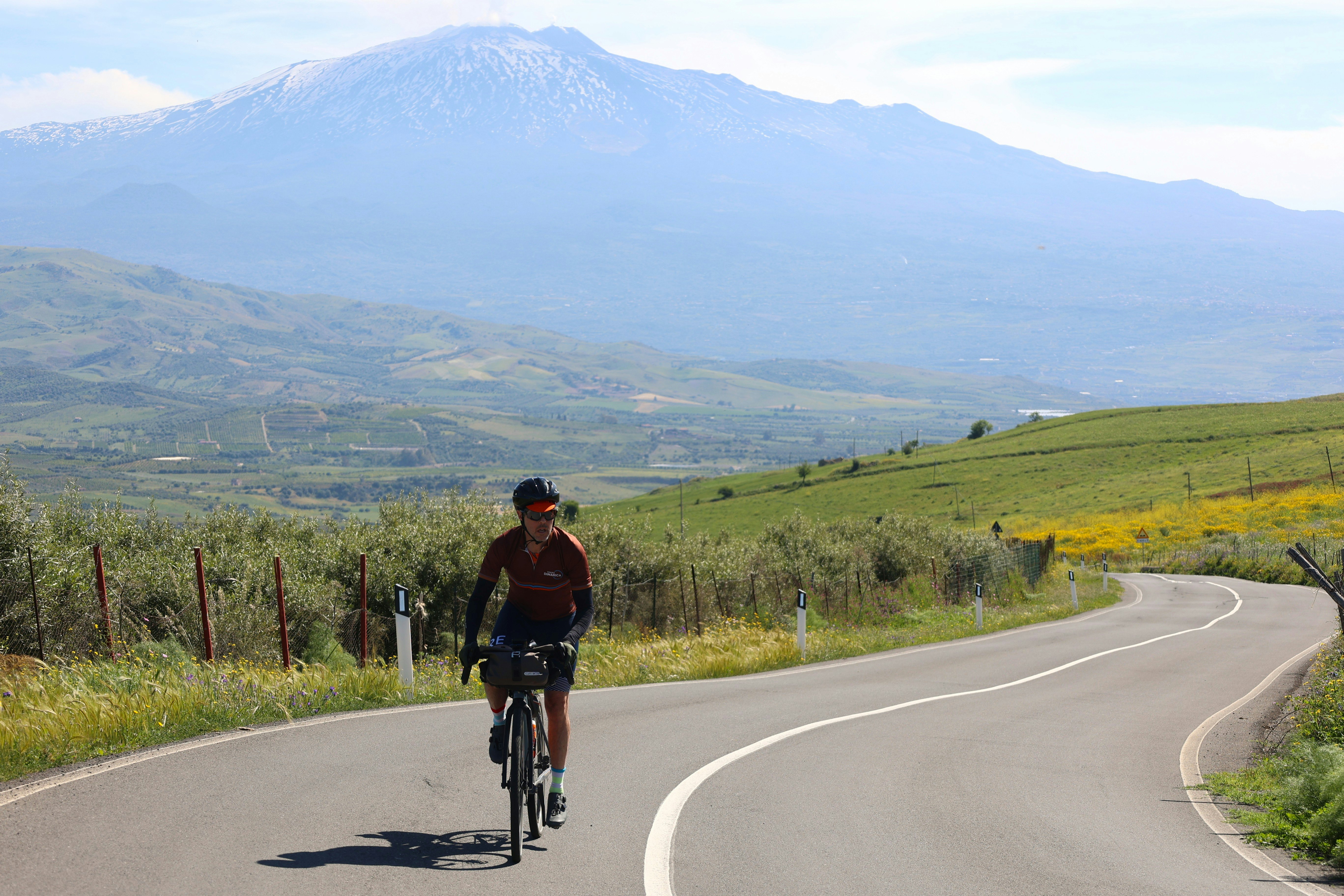 A cyclist pedaling uphill on a steep incline through flower-covered fields in the foothills of a volcano.