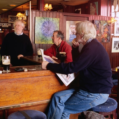 Three men sit at a bar with newspapers and pits of Guinness at the bar in front of them
