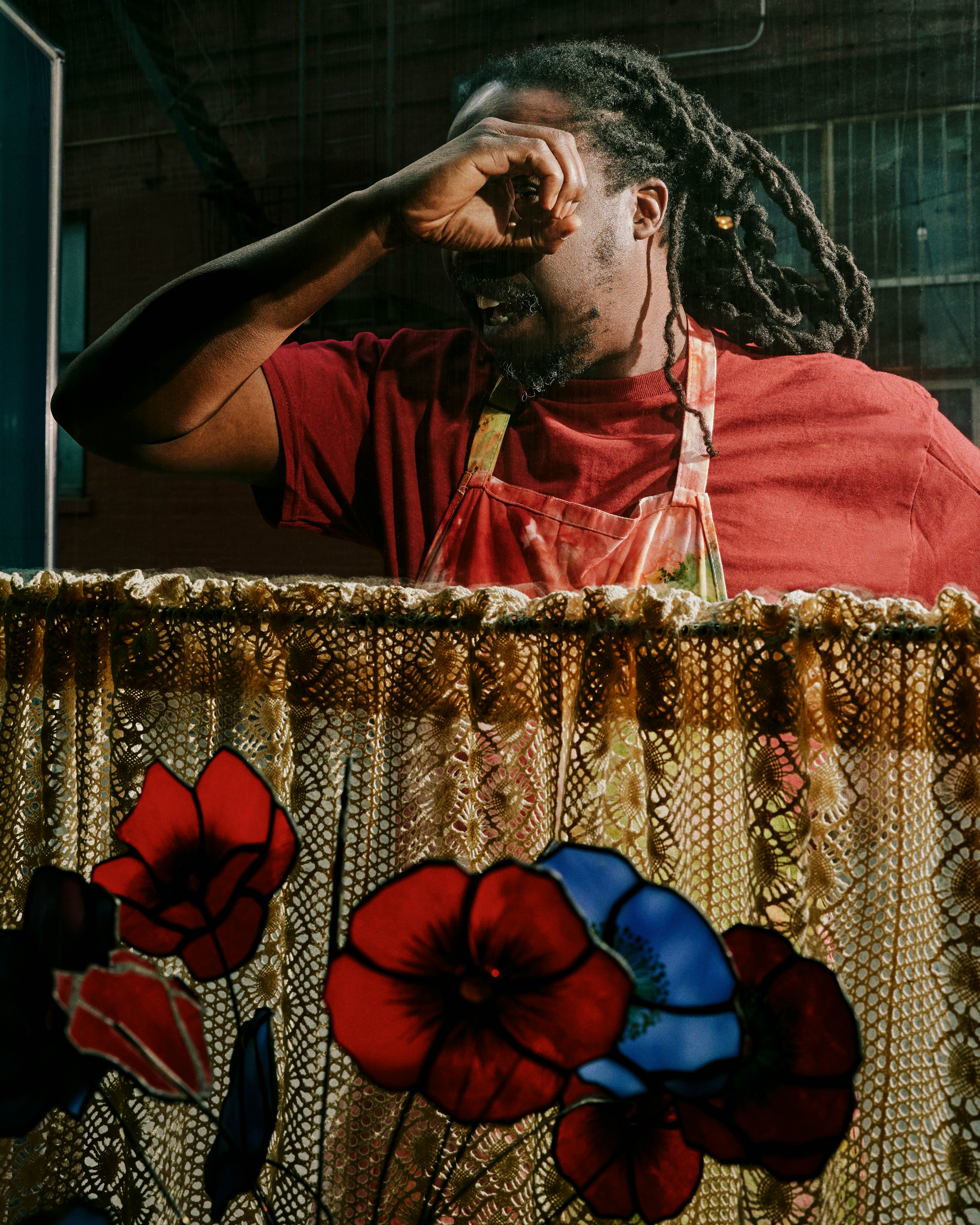 A man in a red shirt peers into a window half covered with a lace curtain and in front of a glass flower arrangement.