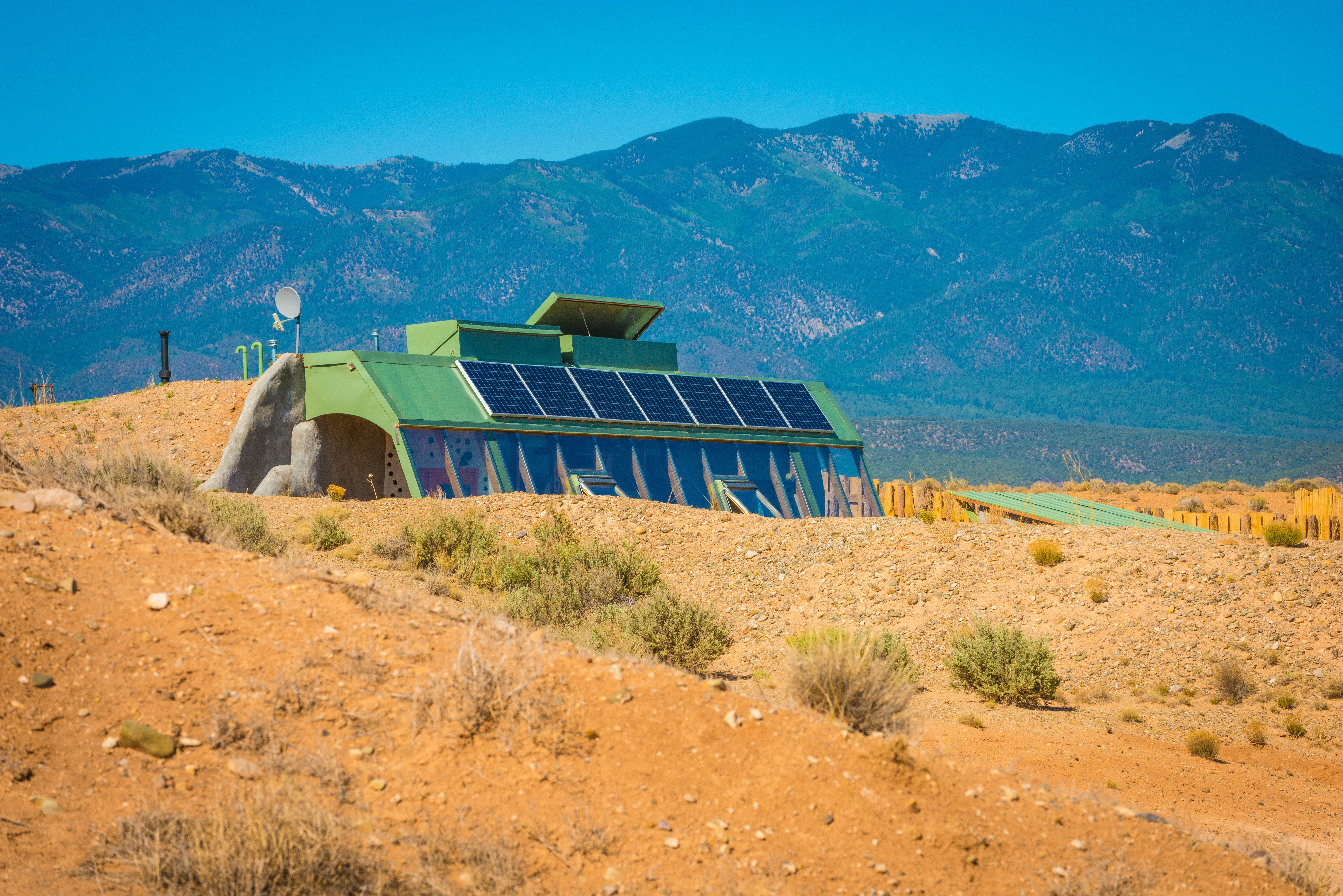 A home built into desert earth covered with solar panels with mountains in the distance on a sunny day.