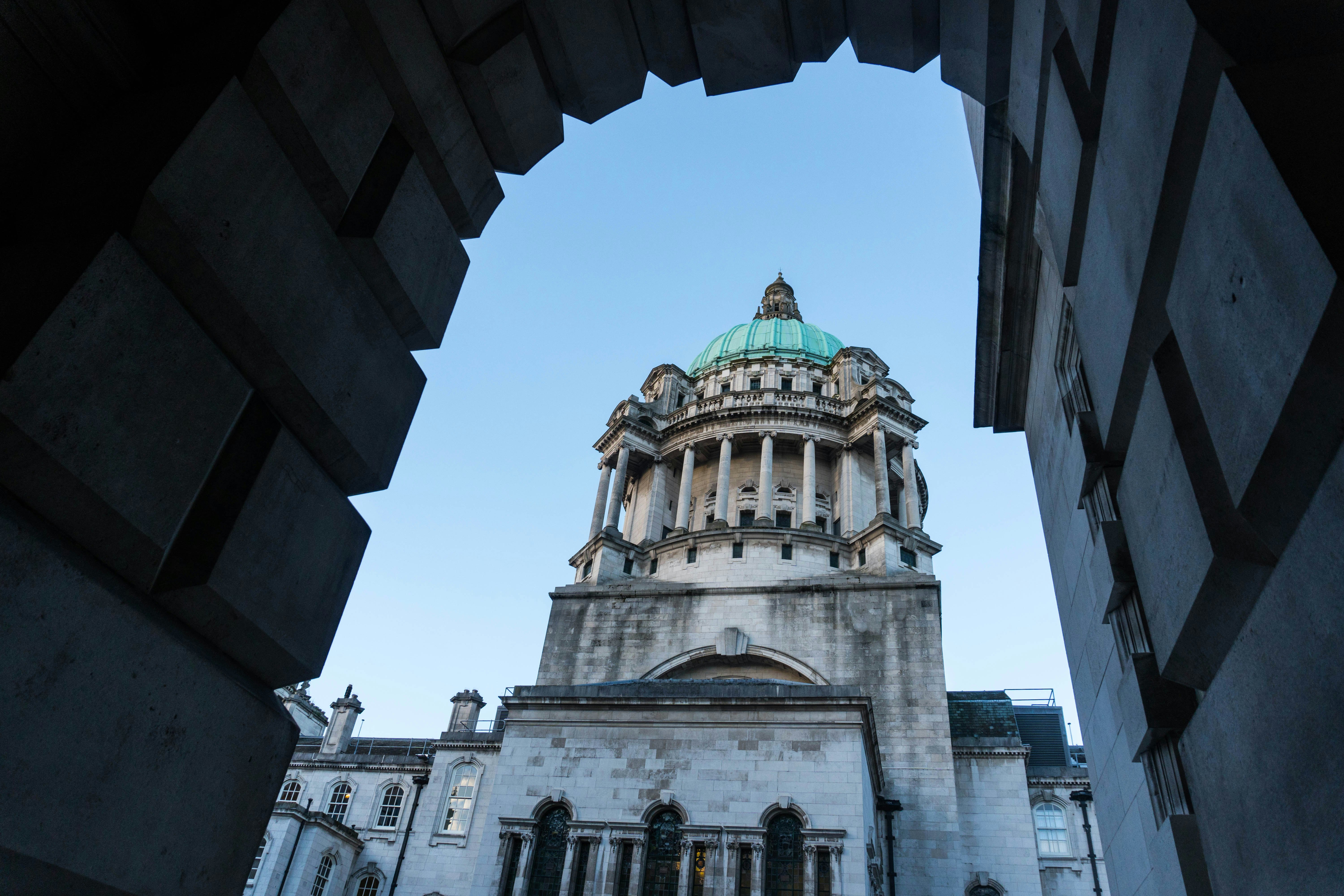 The central stone tower of Belfast's City Hall is visible through an archway against blue sky.