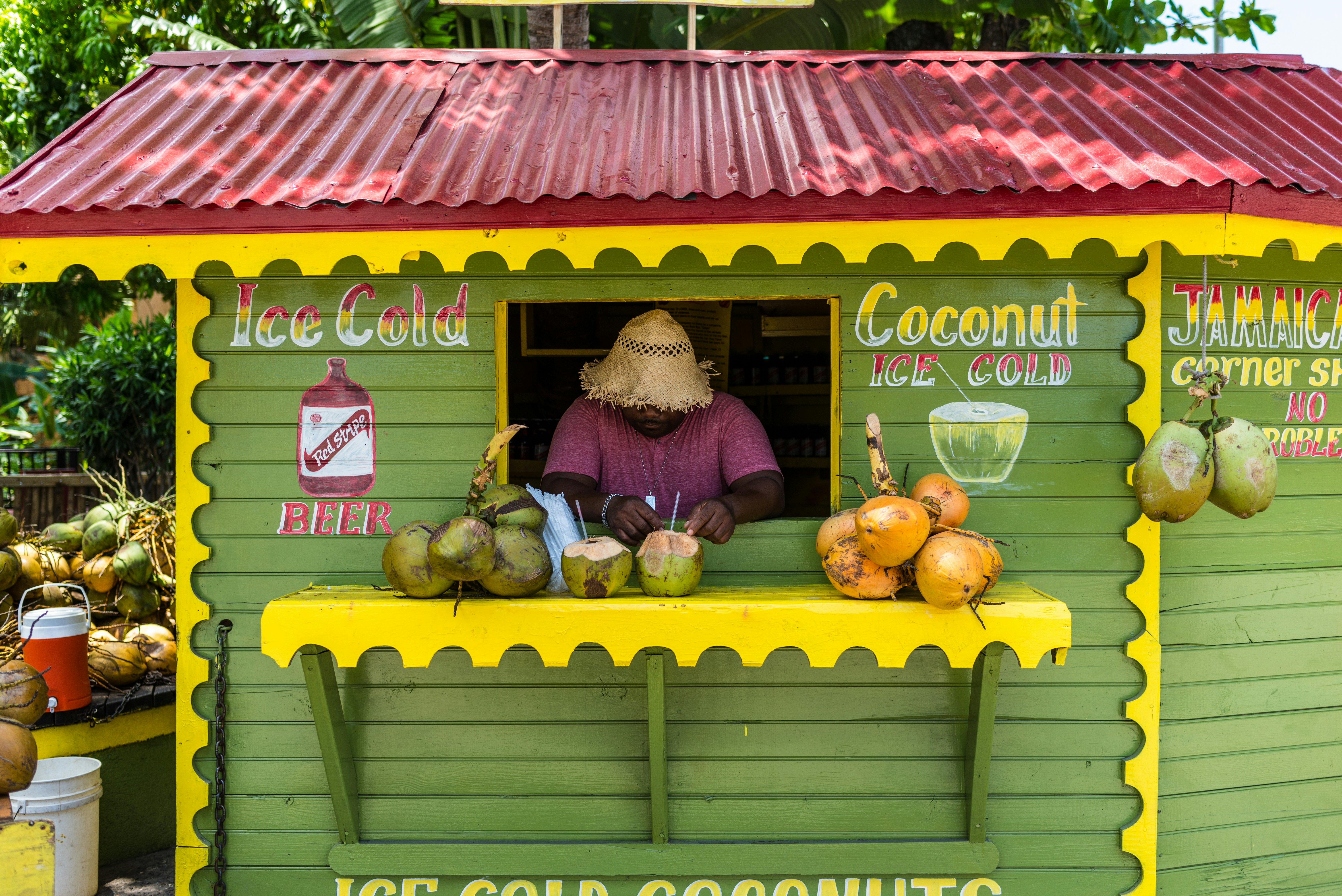 A man wearing a straw hat appears at a window in a green stand advertising beer and coconut drinks; there are coconuts on a yellow shelf in front of the window.