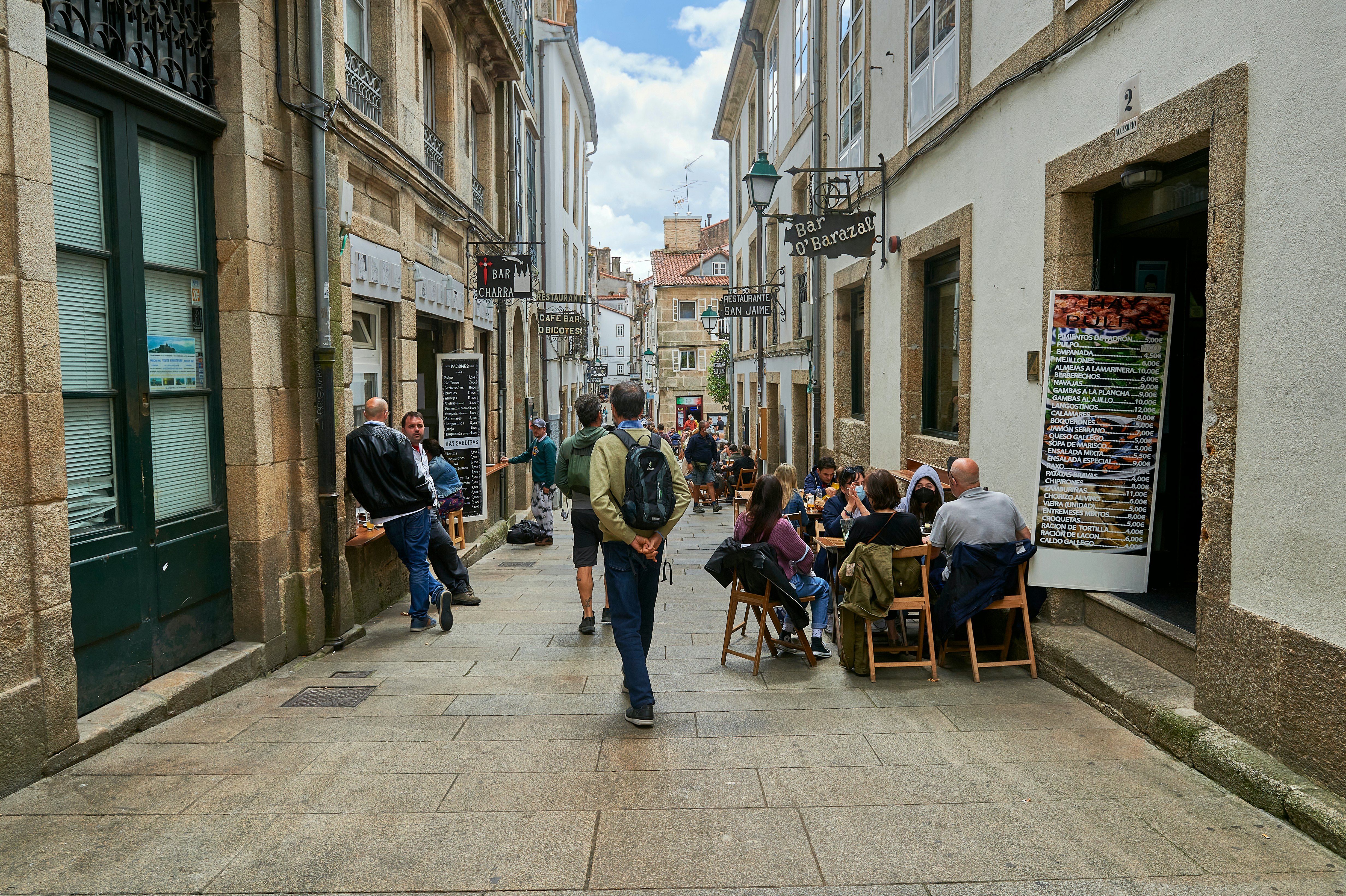 People standing, walking, and sitting in an alleyway between buildings on a sunny day.