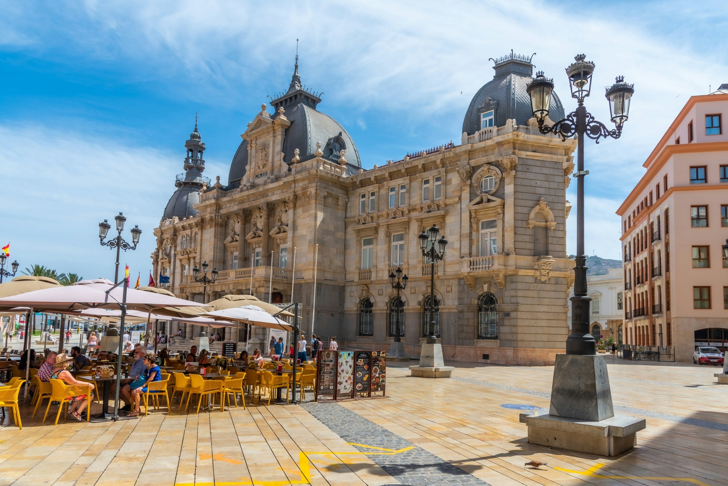 People are strolling and dining in front of Palacio Consistoral in Cartagena, Spain, on a sunny day.