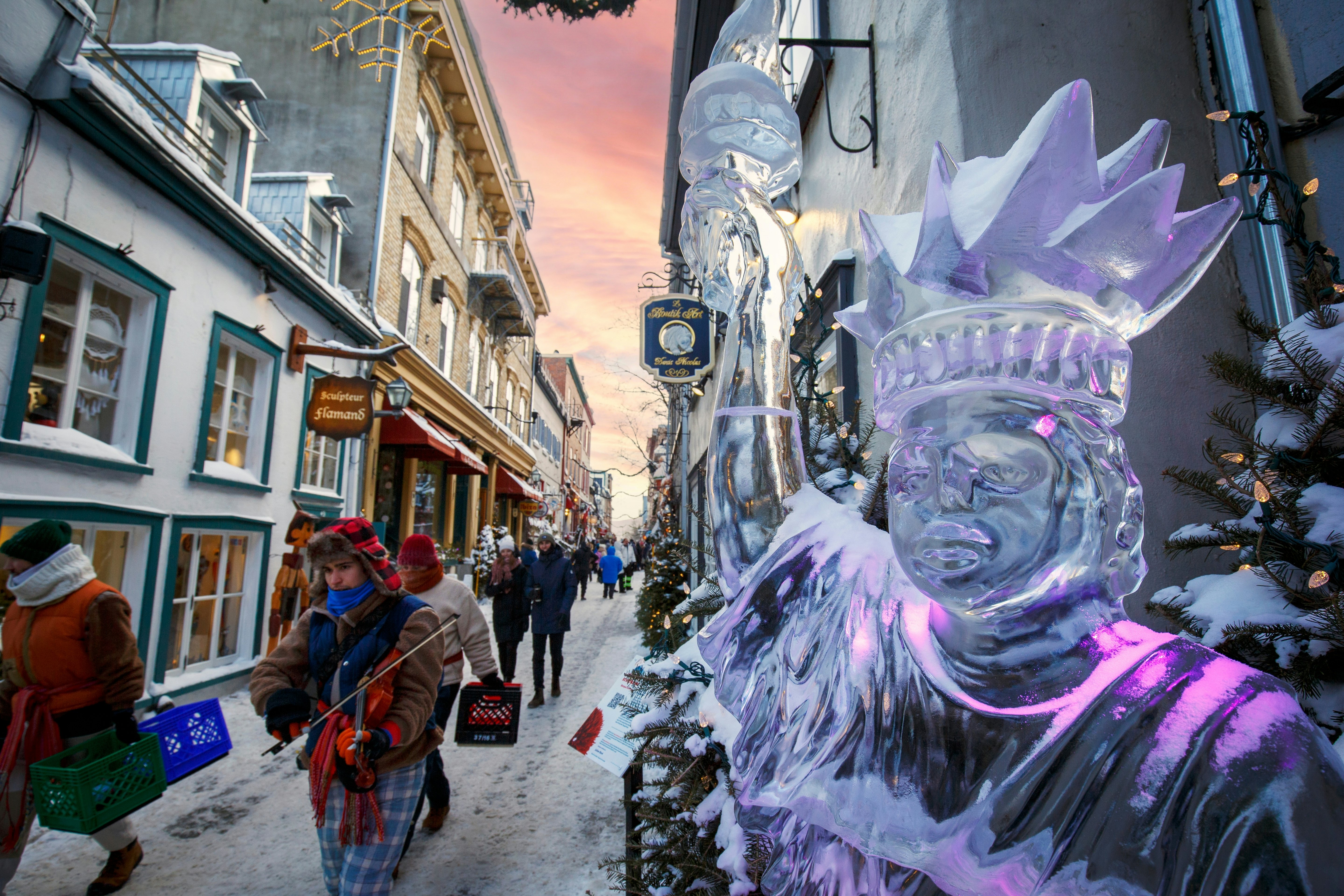 An ice sculpture of a woman holding a flaming torch aloft stands on the edge of a snow-covered street.