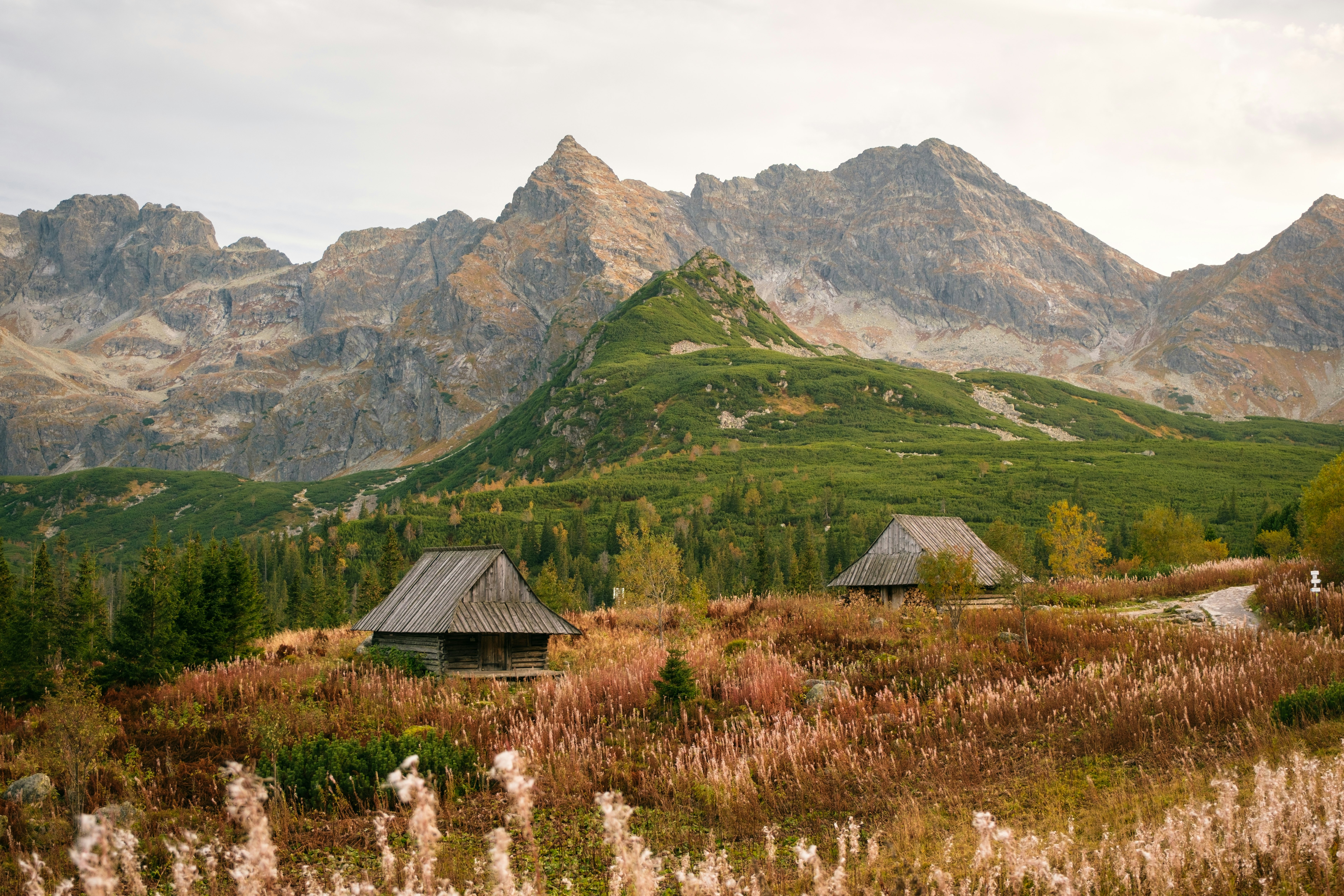 Two huts in a dry grassy field; a green hill and rocky mountains are behind them.