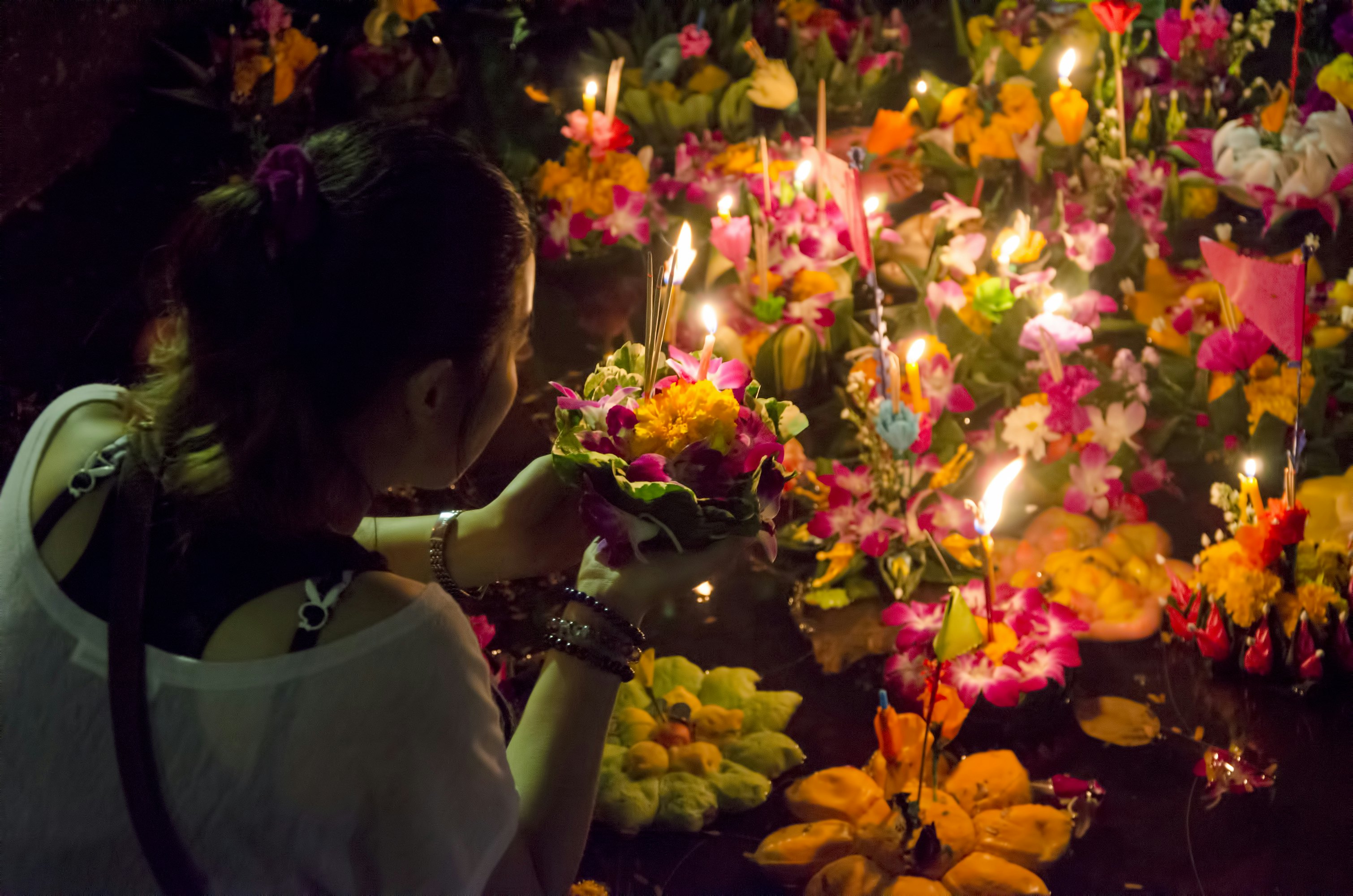 Woman puts flowers with candles stuck in them in a body of water with other similar flower and candle bundles.