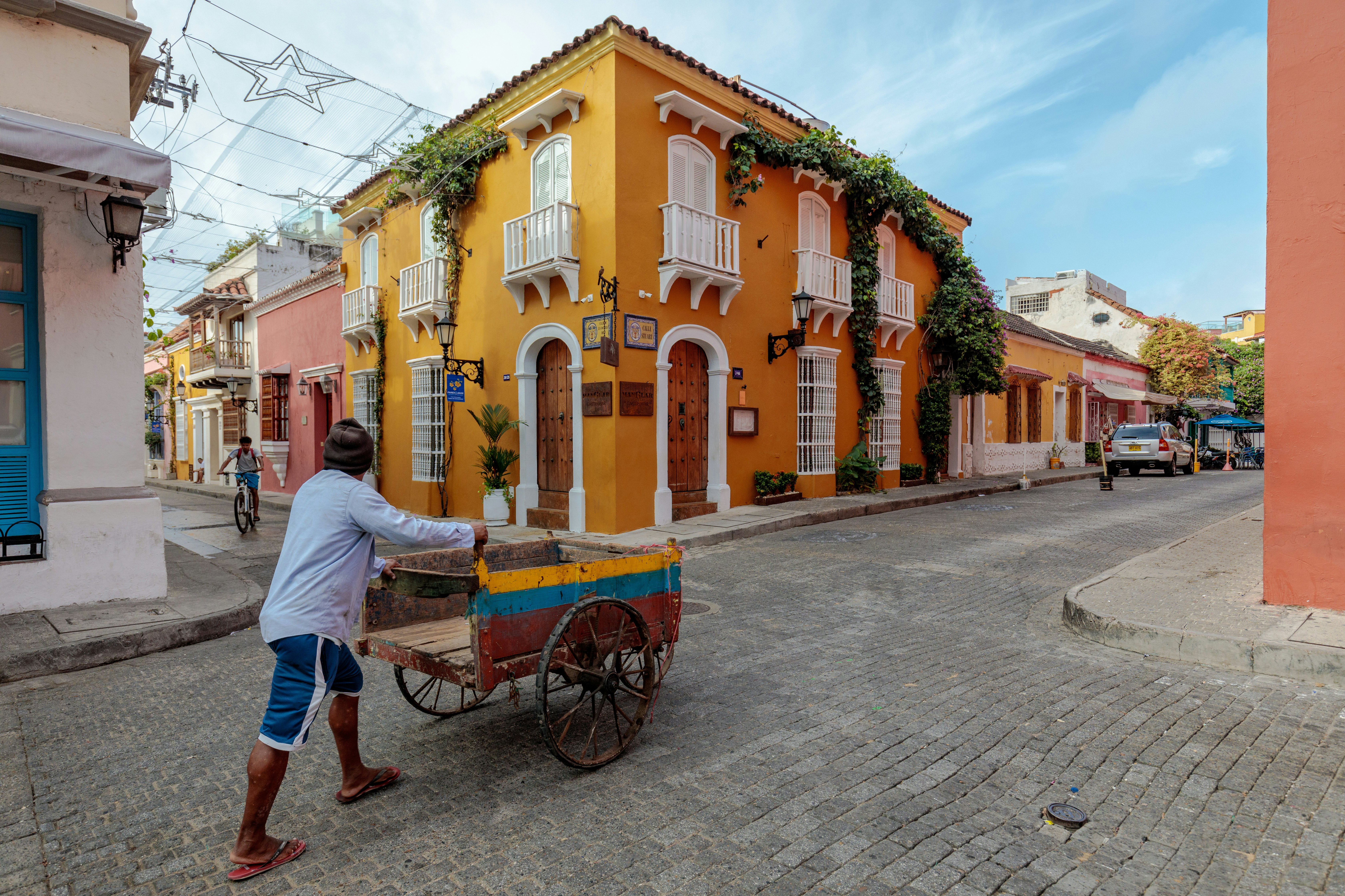 A man pushing a cart walks by colorful buildings on a cobblestone street