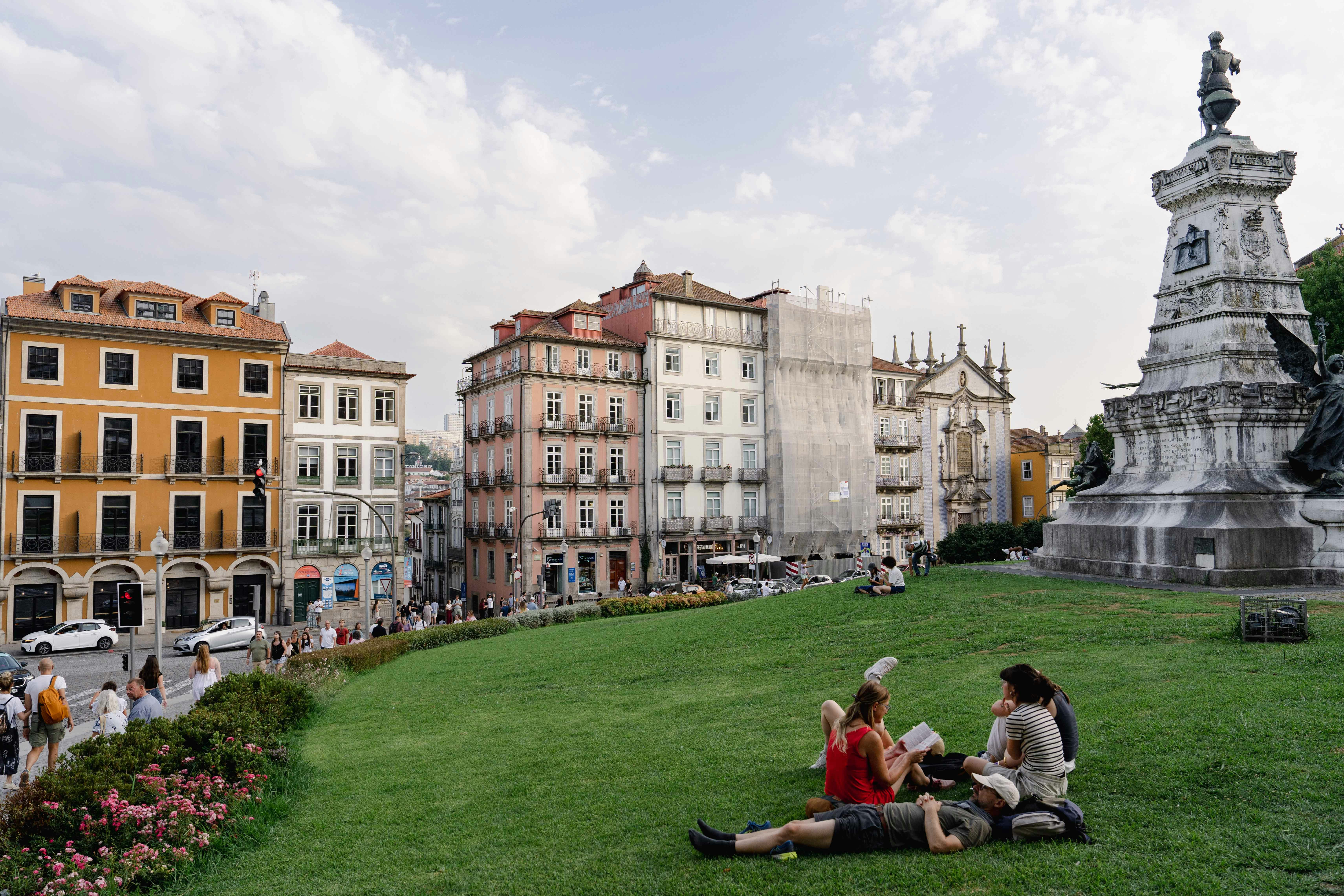A group of people are sitting and reclining on the green grass of a park in Porto, Portugal; pastel-colored historic buildings are on the opposite side of a street in the background.