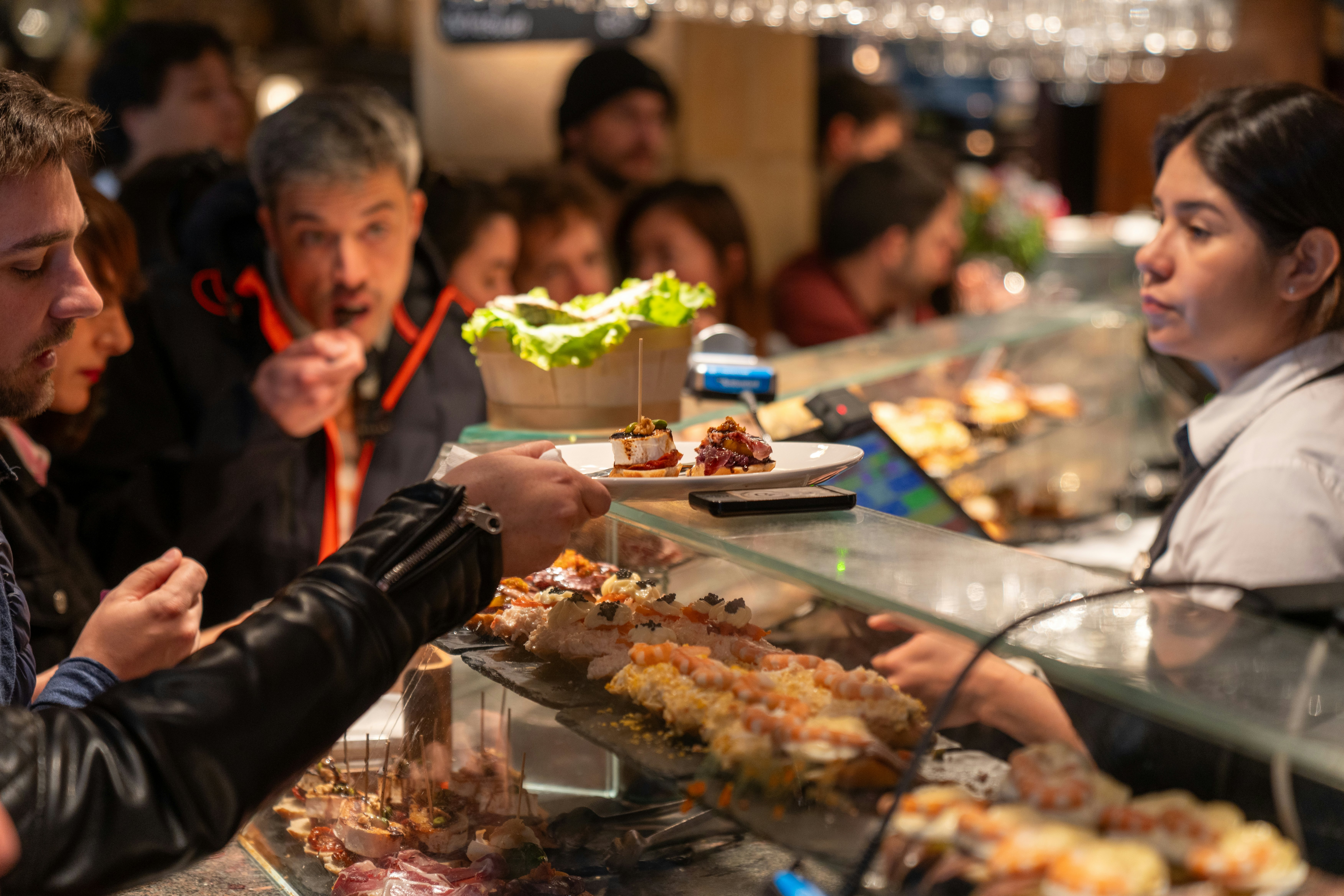 People at a bar with food displayed on shelves. One patron takes a plate from a glass shelf, with a server behind the counter.