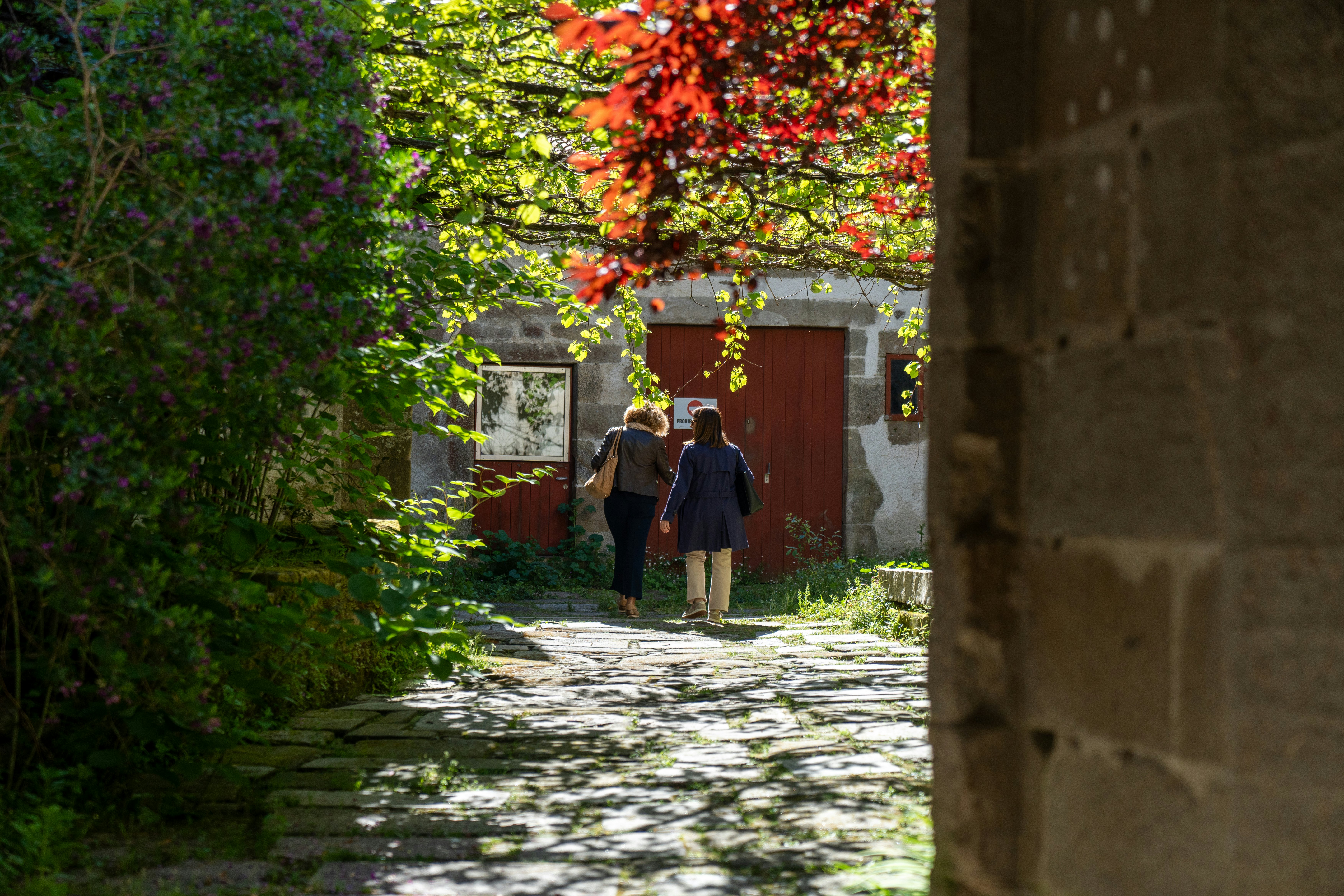 Two people walking on a cobblestone path toward a stone building with red door on a sunny day.