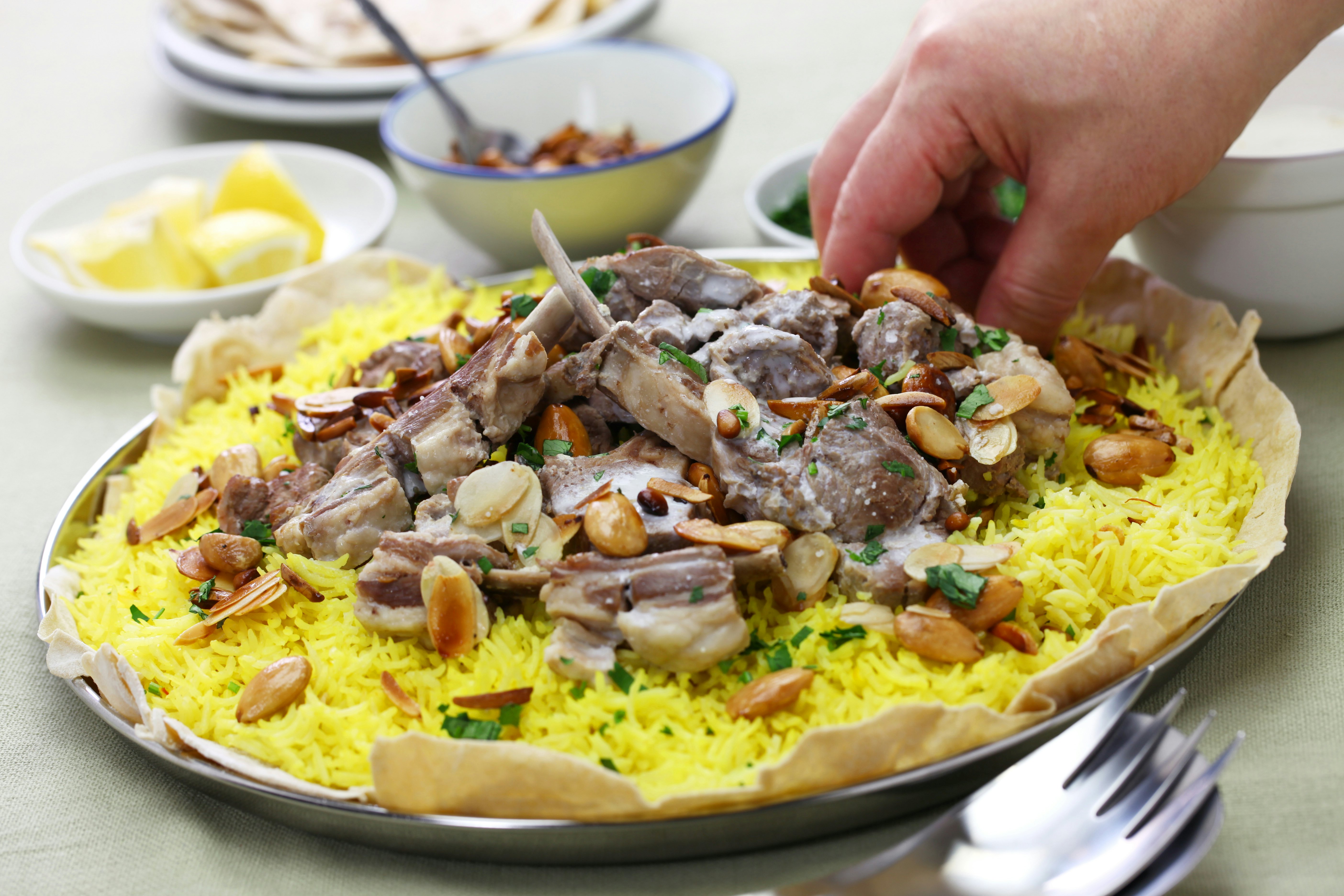 A hand reaches towards a large rice dish topped with lamb, pine nuts and herbs.