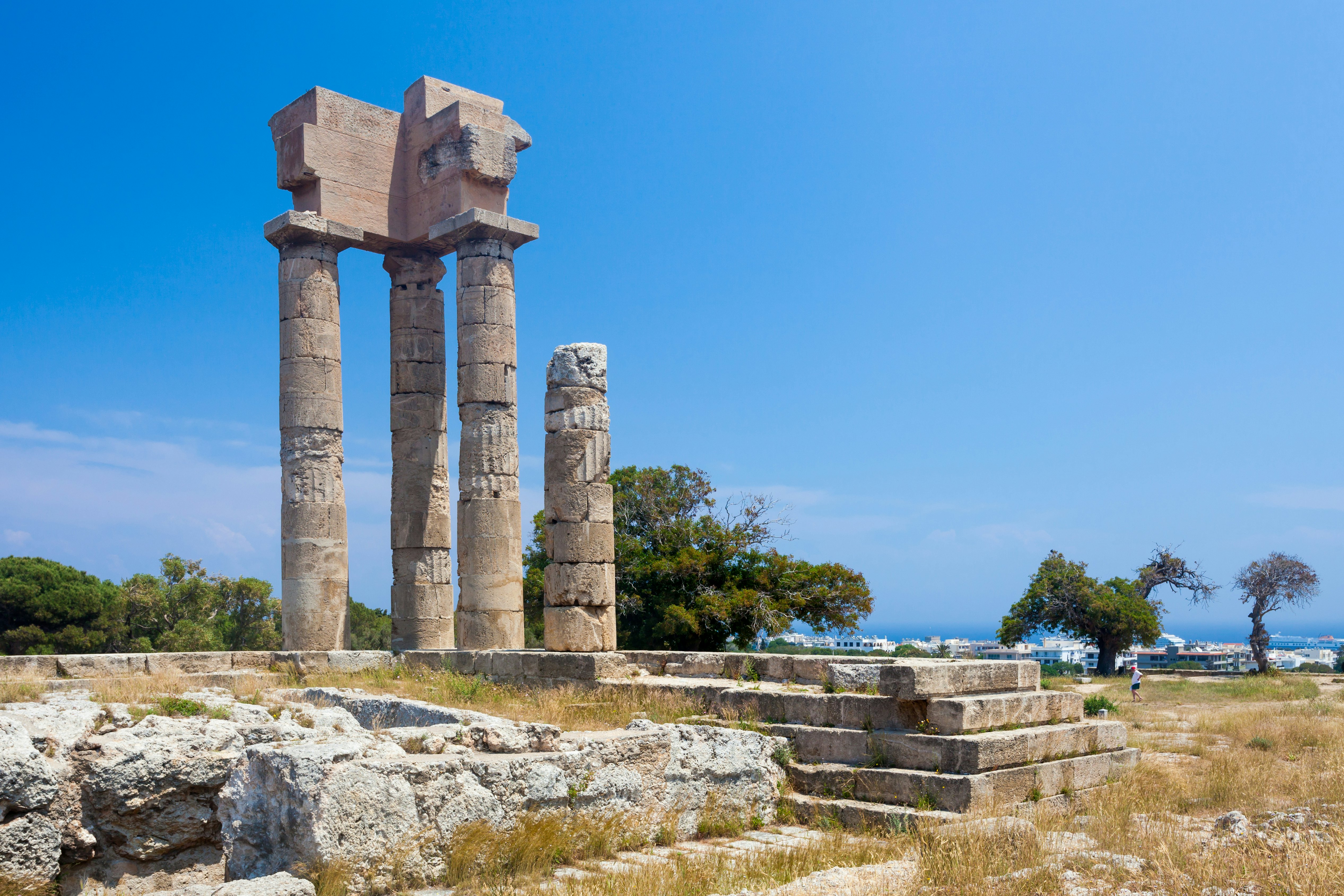 A cluster of columns and other ruins are seen at an archeological site, with the blue sky above and the sea visible in the distance.