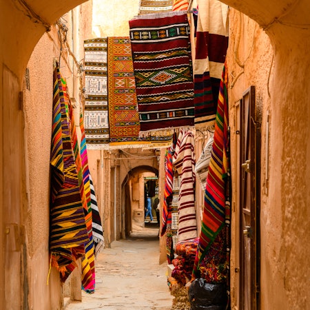 Colorful fabrics are displayed on walls seen through an archway in an Algerian town.