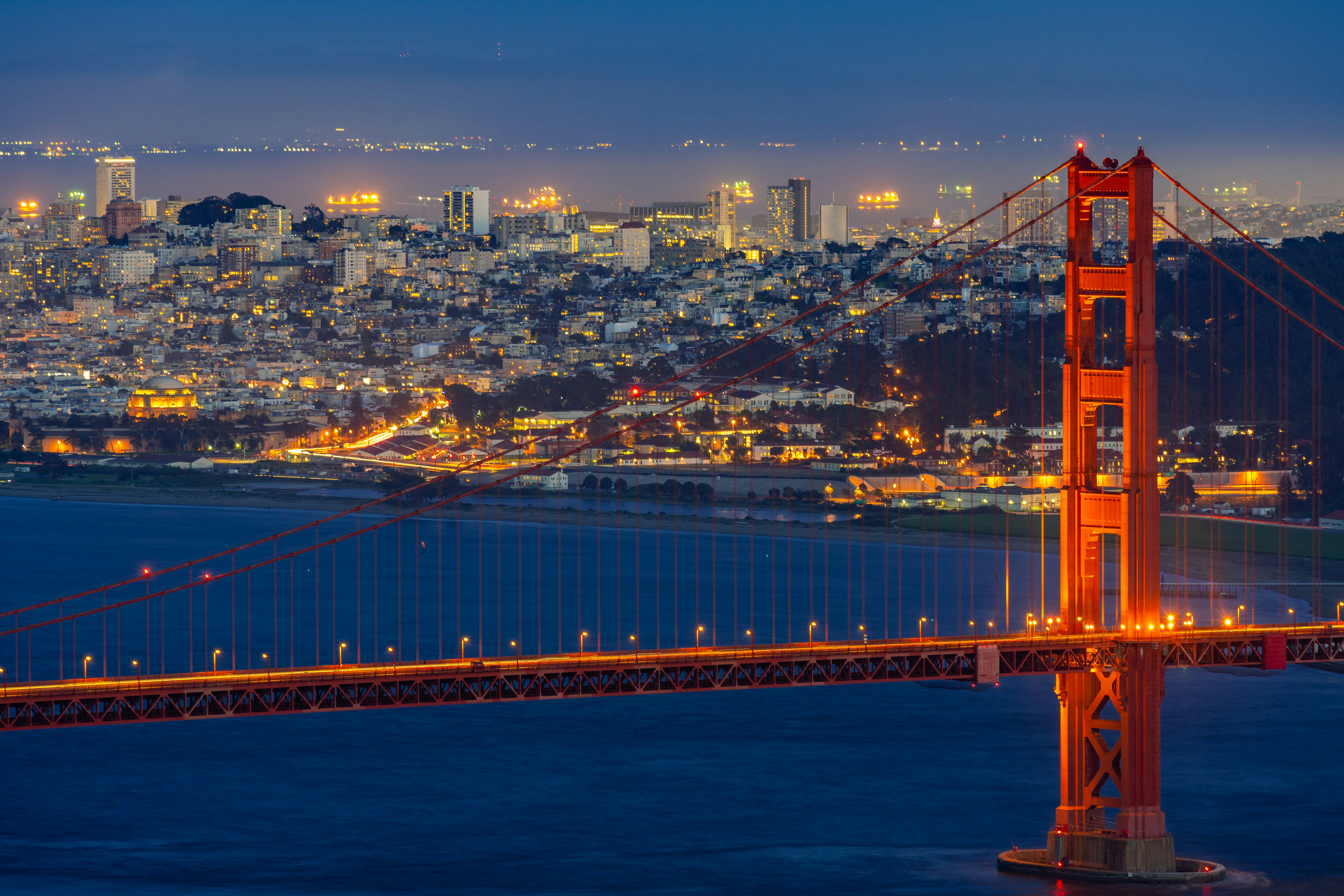 The illuminated Golden Gate Bridge at night over deep blue water with city buildings and homes in the background