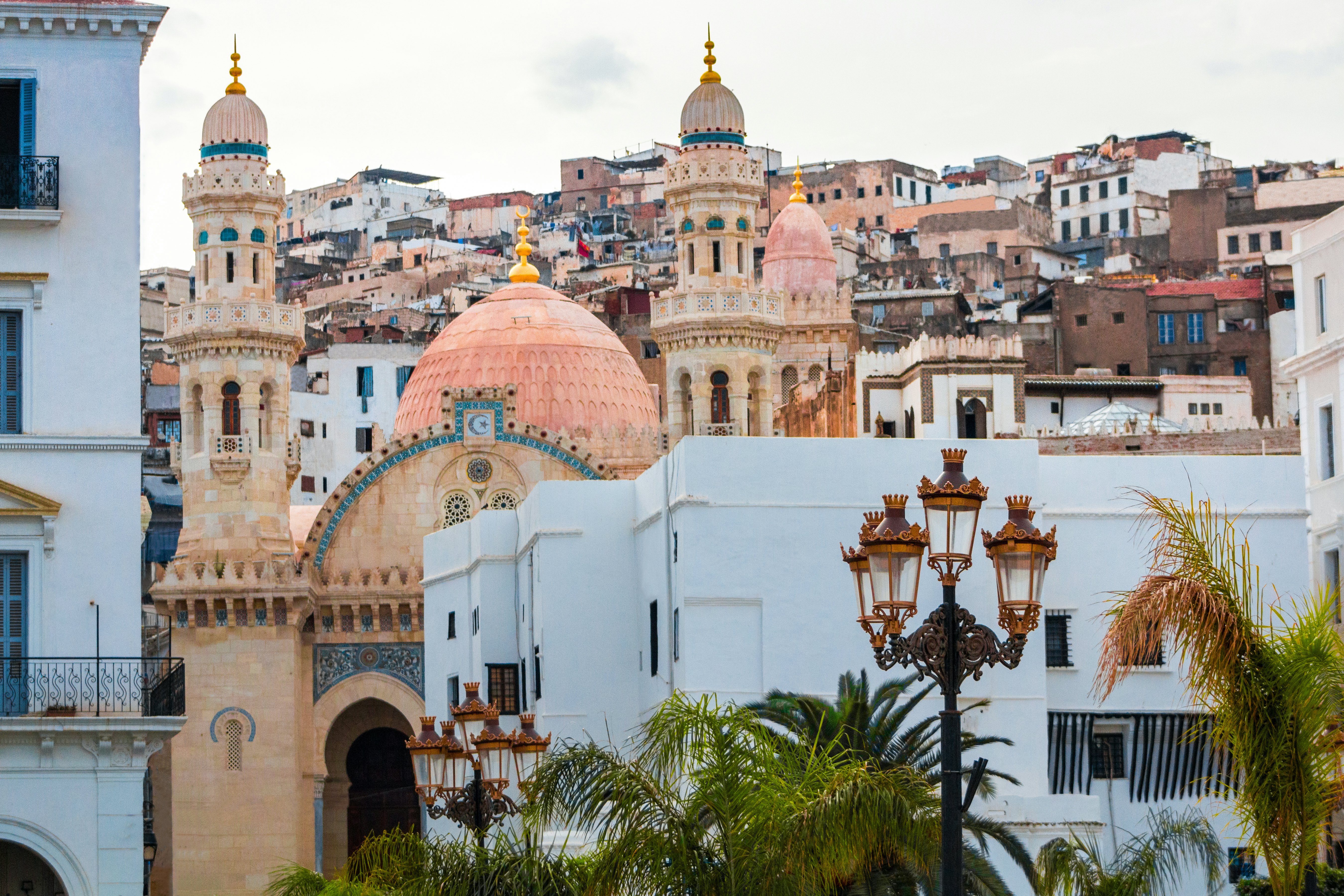 The towers of a mosque in Algiers appear in the foreground, with a hillside of historic buildings rising behind it.