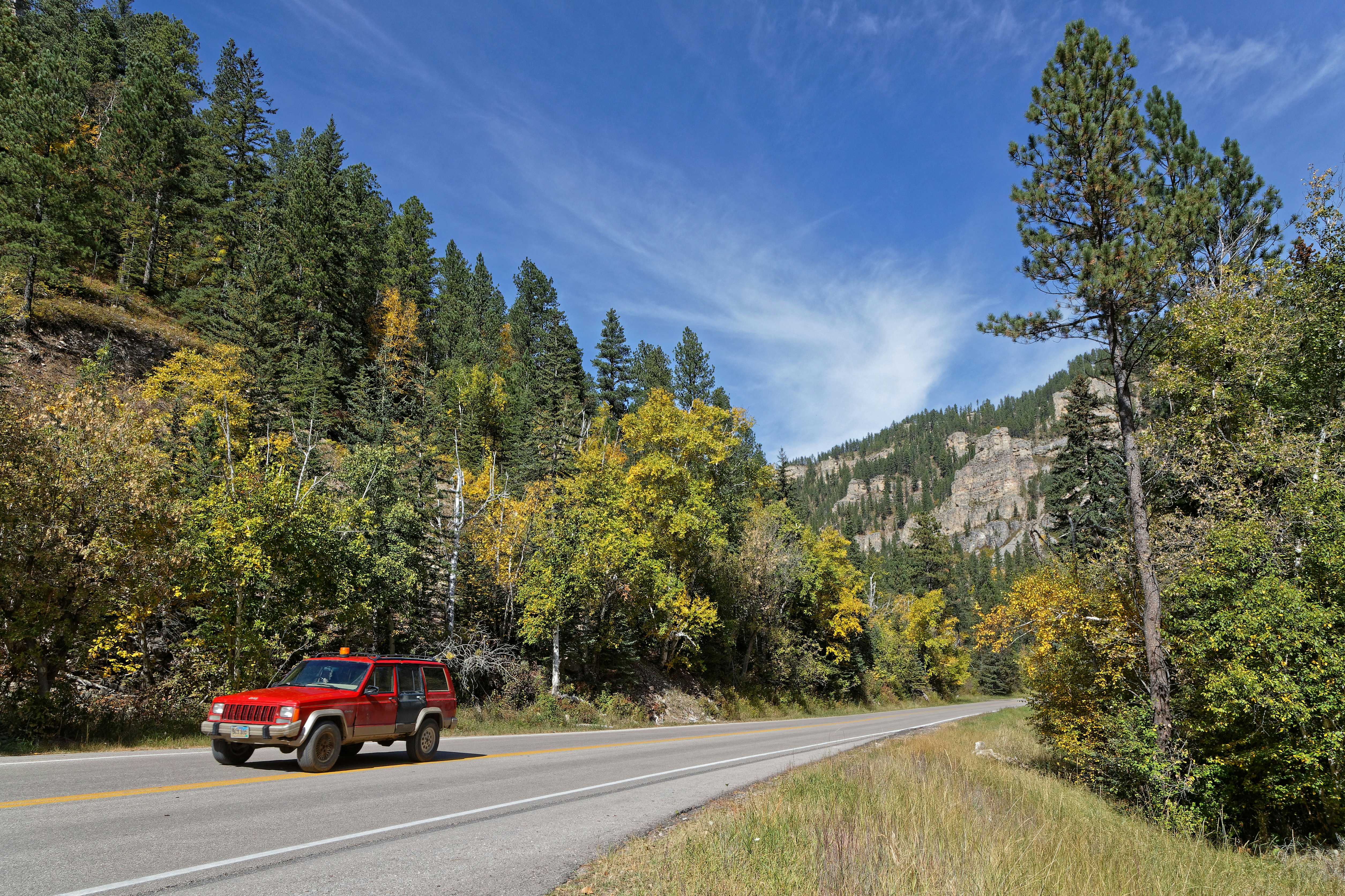 A red car drives along a road in early fall as the leaves on the trees are starting to turn from green to yellow.