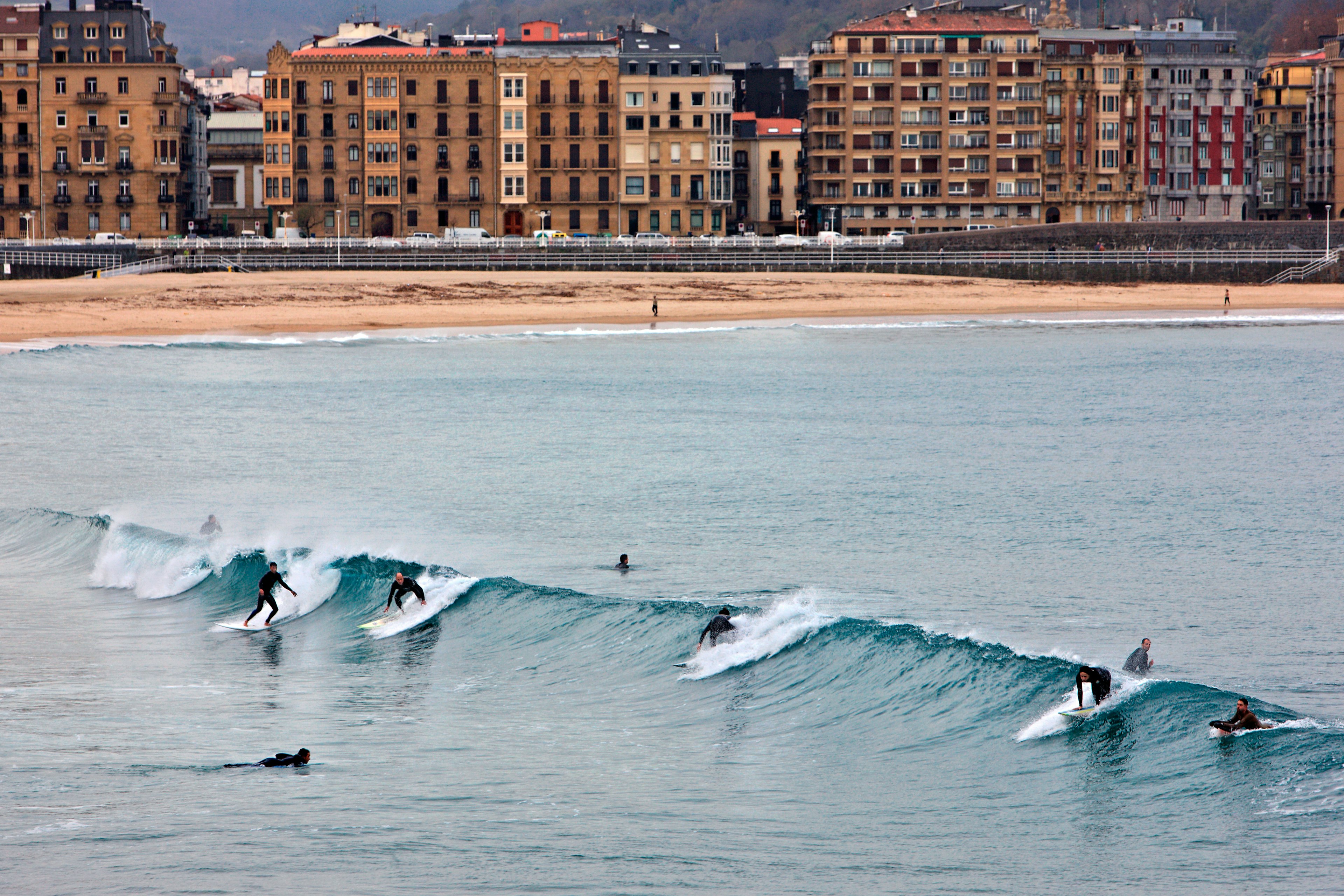 A line of surfers in wetsuits riding a wave off the shore of a golden sand beach; buildings line the road on the other side of the beach.
