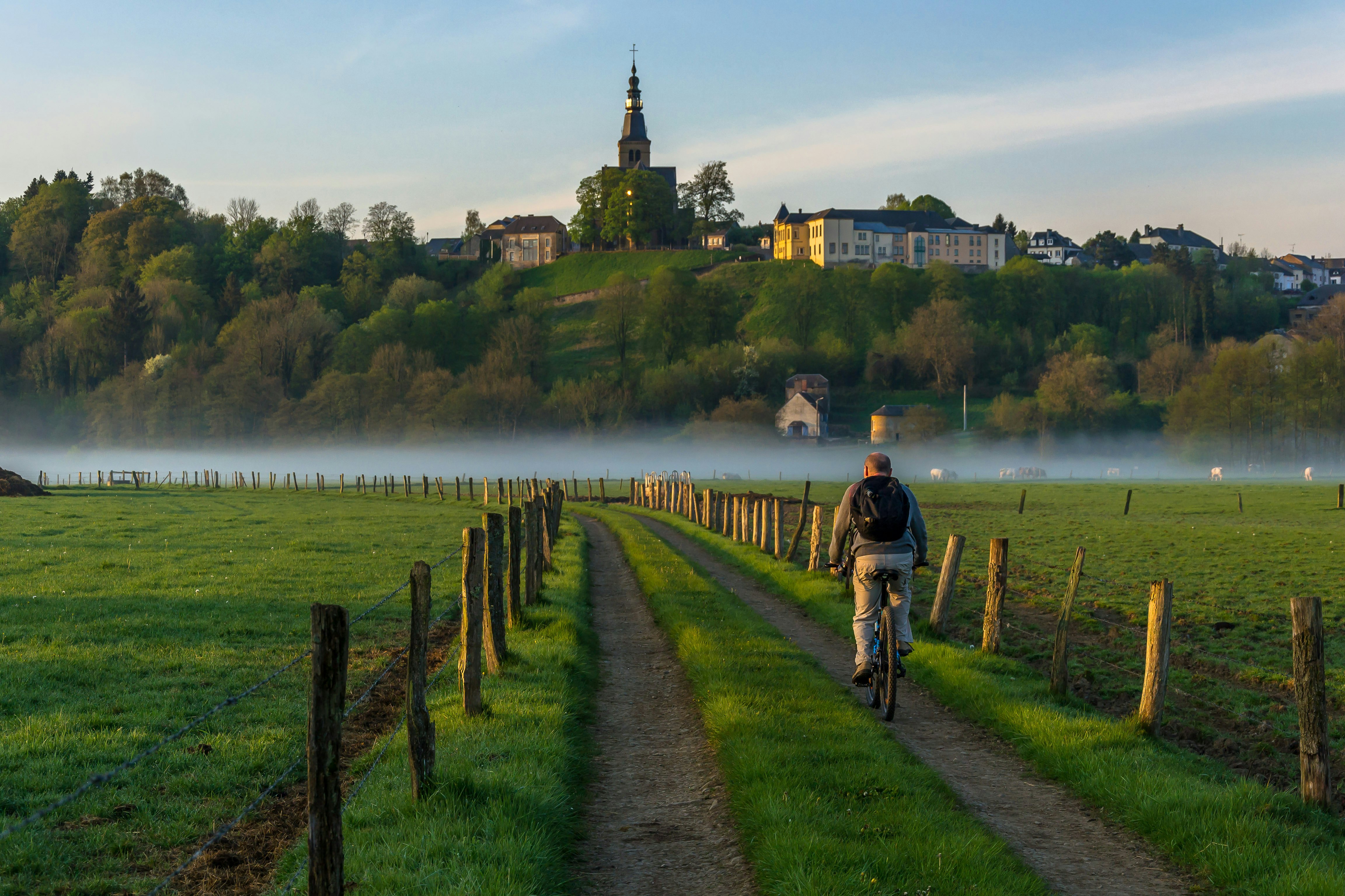 A man on a bicycle is pictured from behind as he pedals down a rural road past fields and toward a misty river. A church and houses are seen on a ridge in the distance.