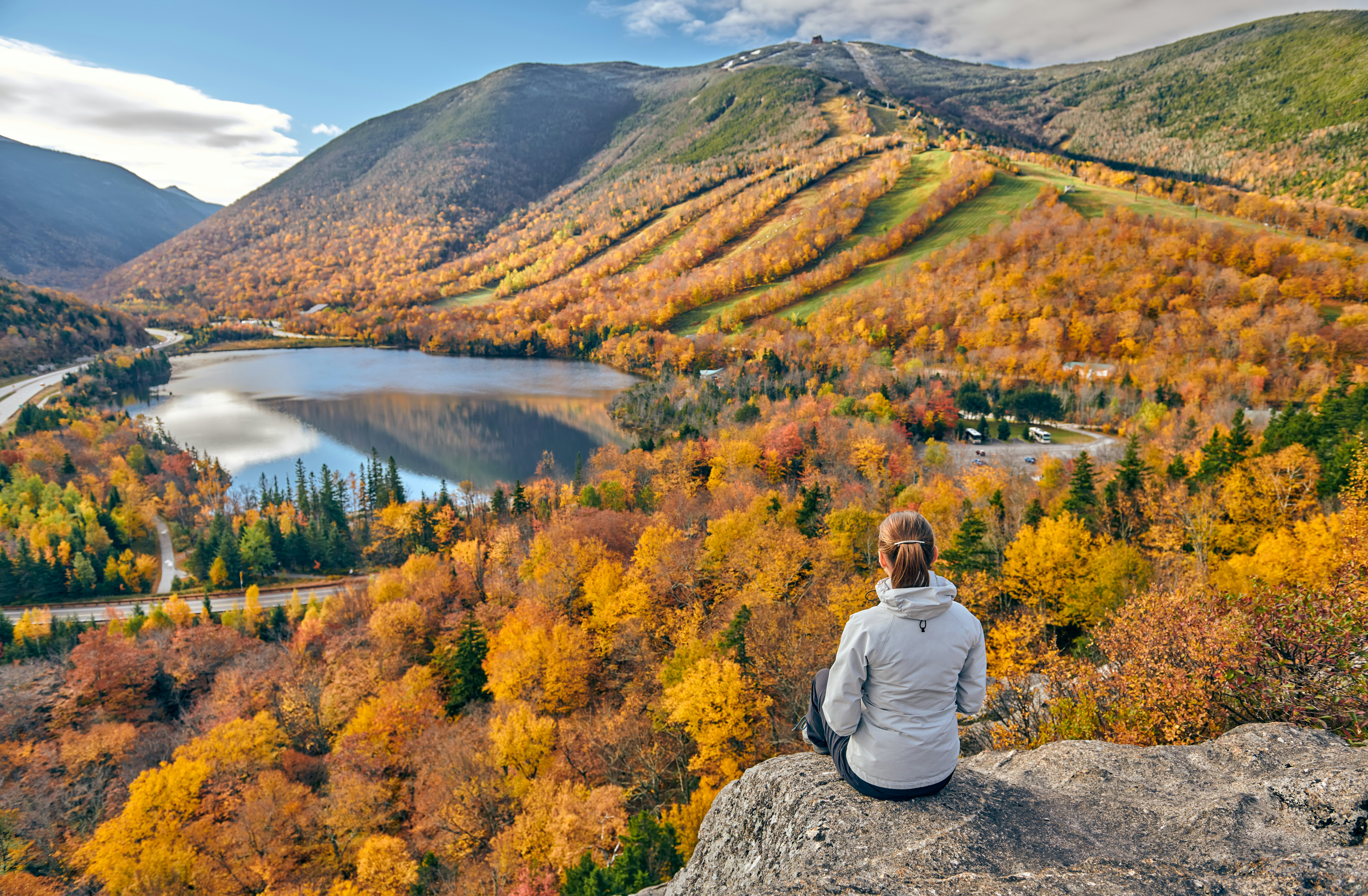 A woman sits on a rock at a viewpoint looking down over woodland in the golden colors of fall.
