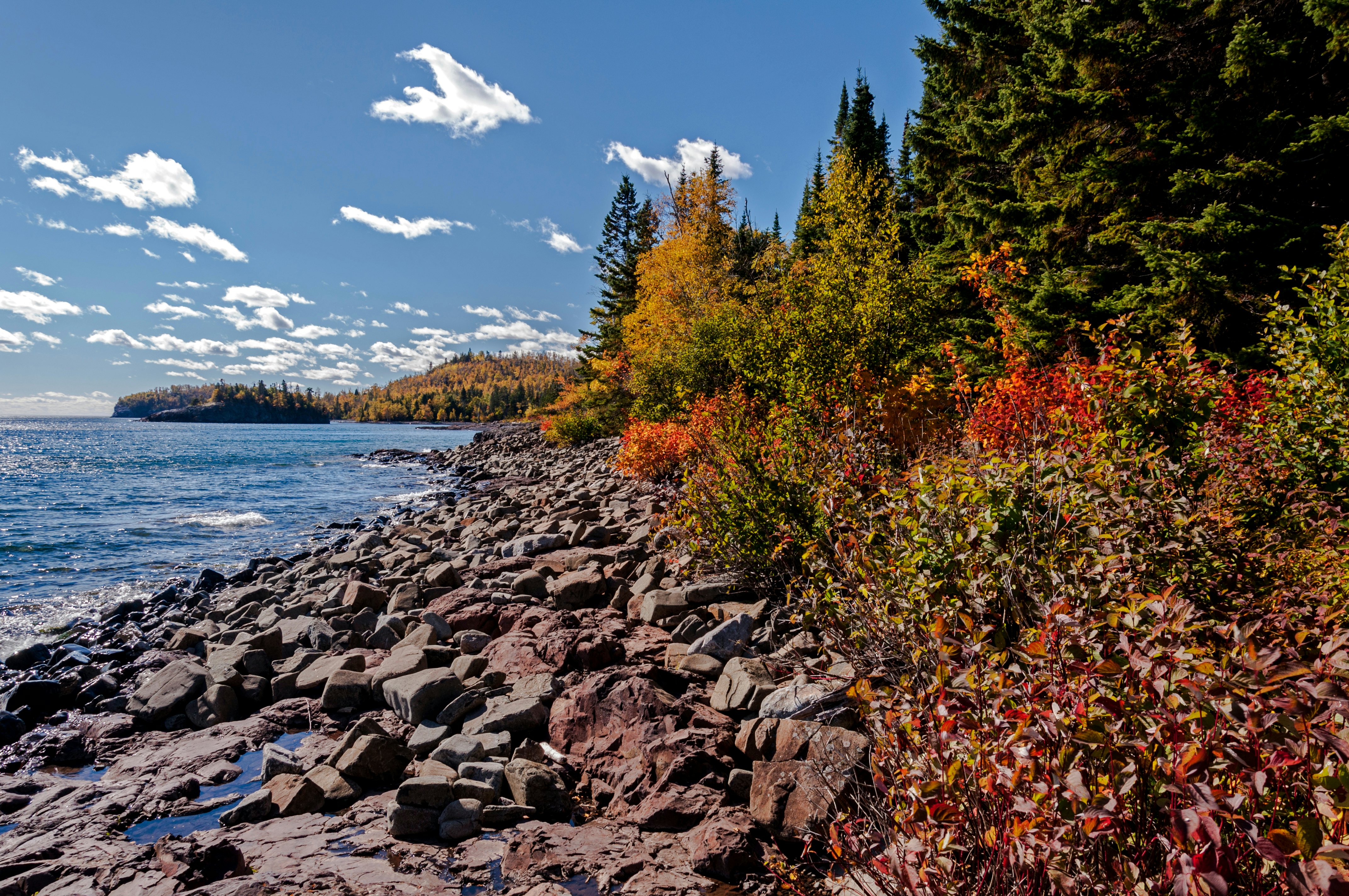 A rocky lakeshore backed by trees glowing orange and red in the autumn sun.