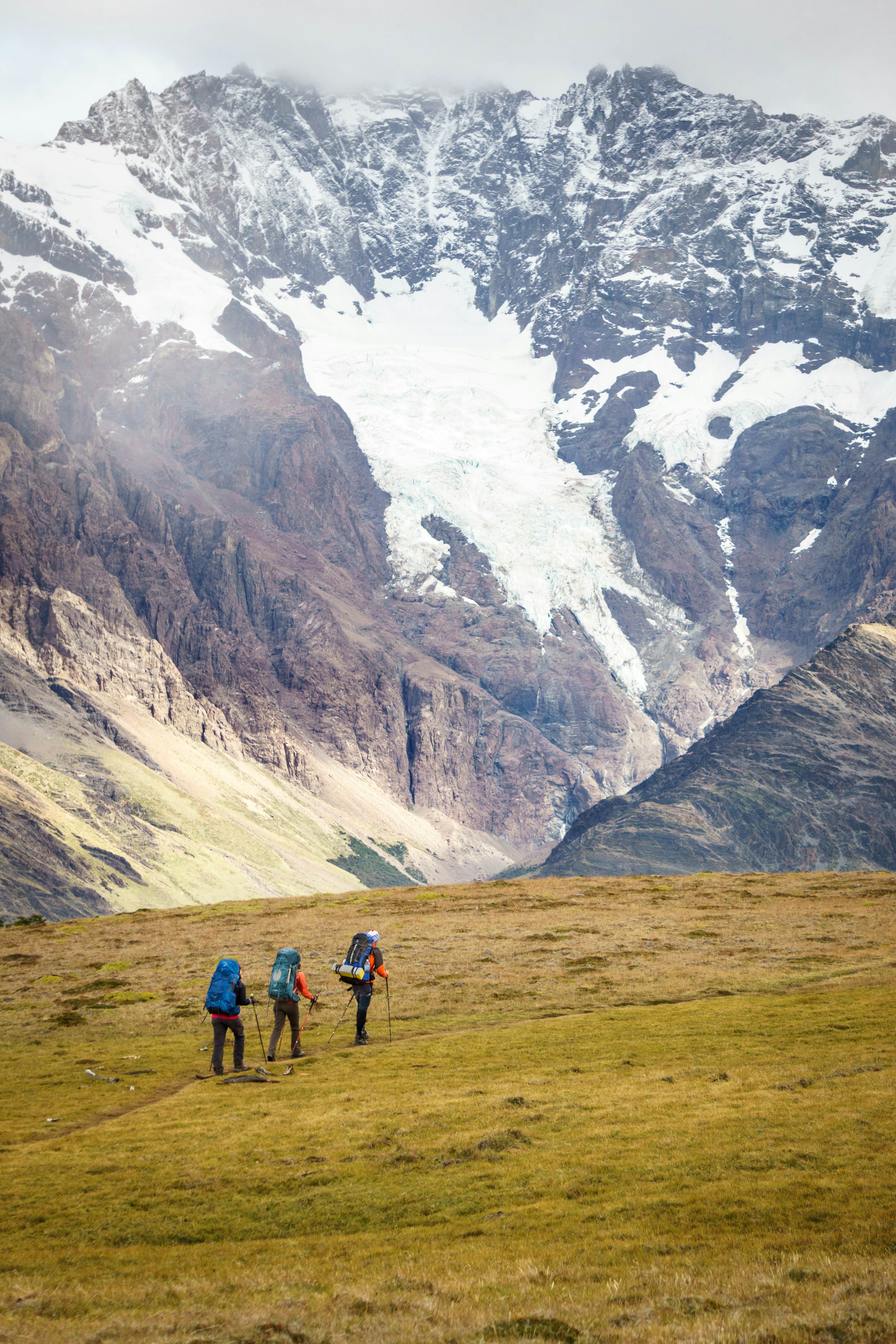 Three hikers walk over a grassy path; patches of snow on a rocky mountain are in the background.