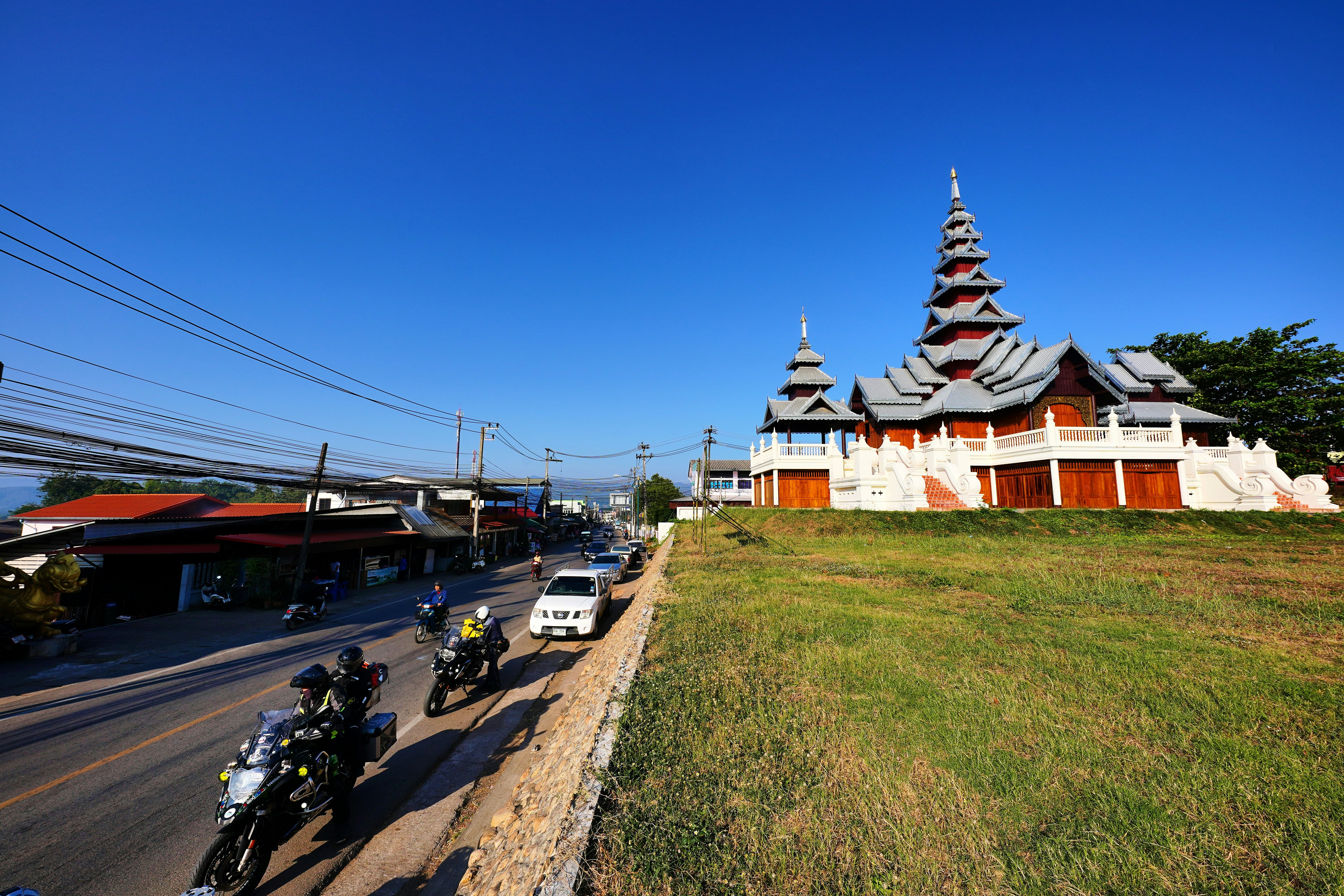 Motorbikes and cars line the road adjacent to a building with white railings and a towering roof