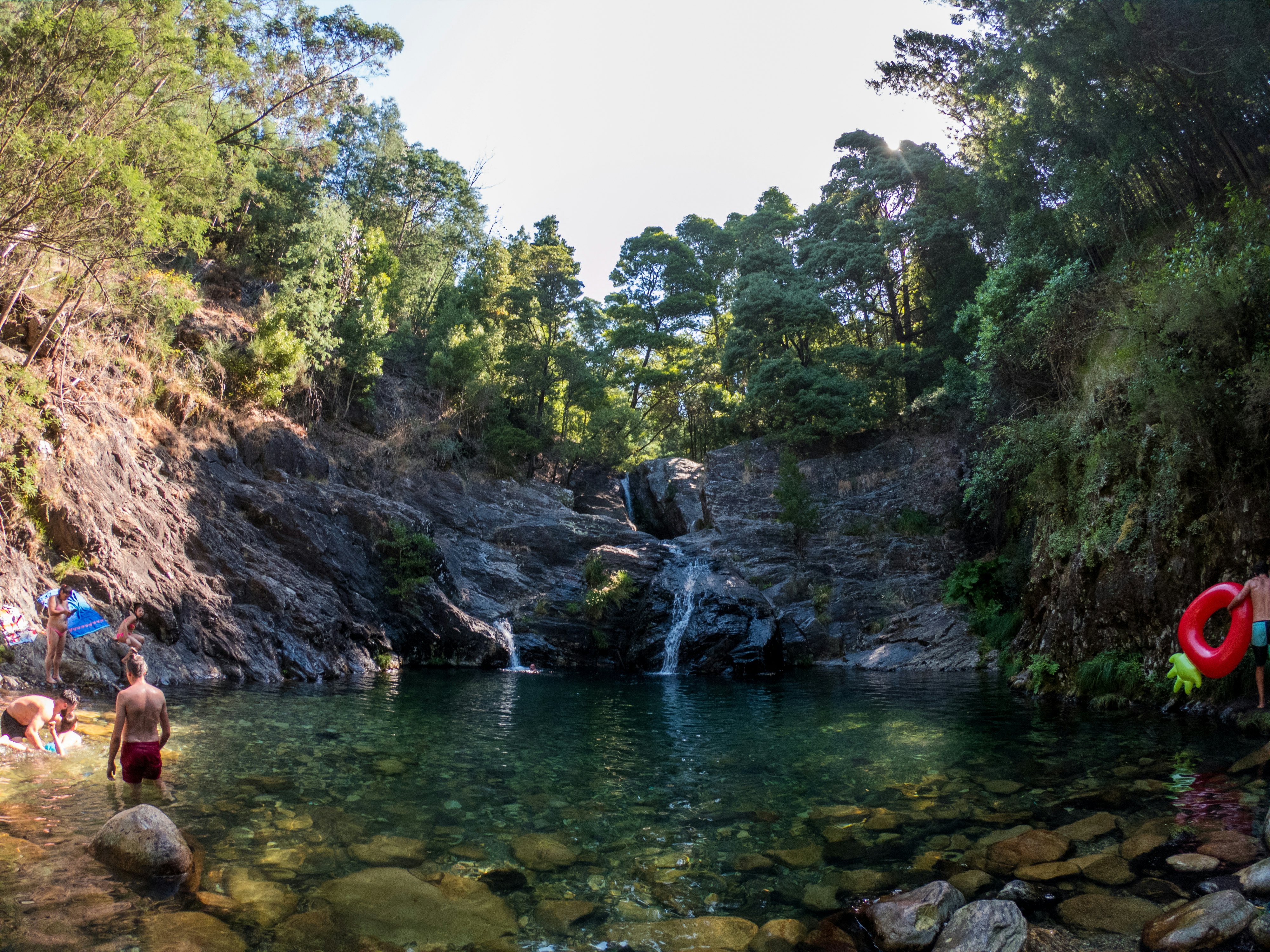 People swim in the lagoon formed by a waterfall, shaded by trees.
