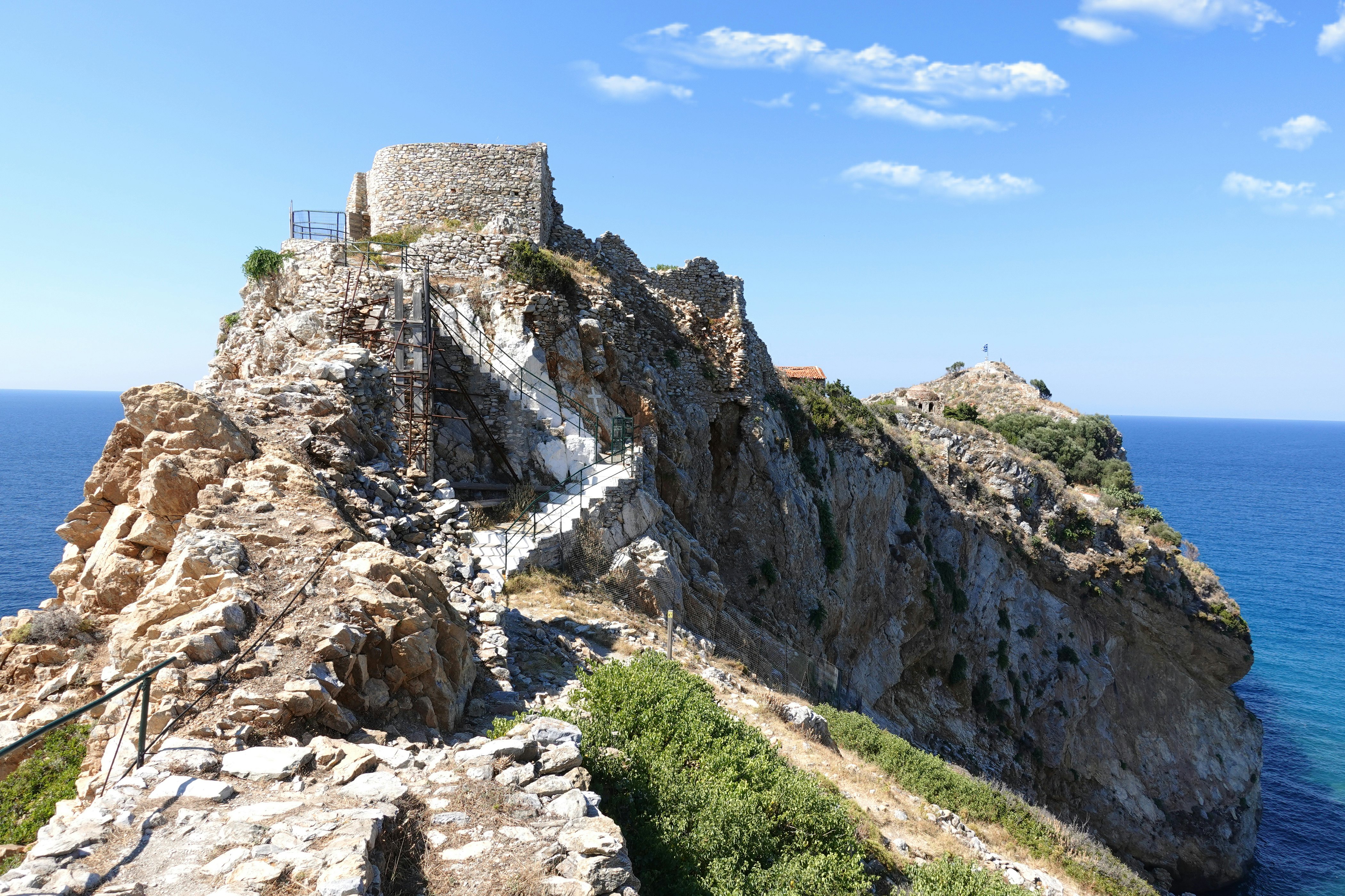 A path leads to ruins on a headland with cliffs going down to the blue sea.
