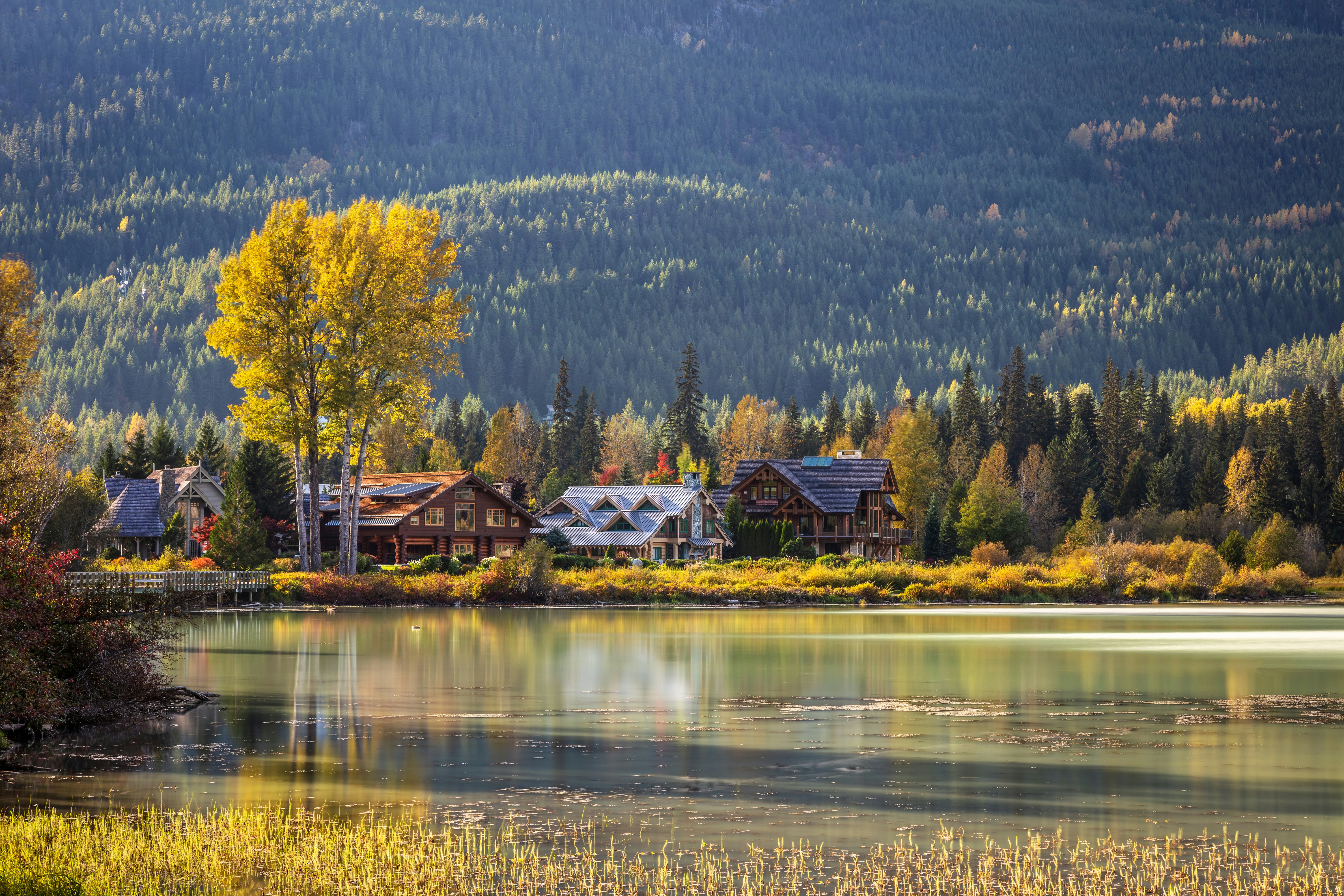Homes by the shore of a greenish lake at the base of a tree-covered hill in autumn.