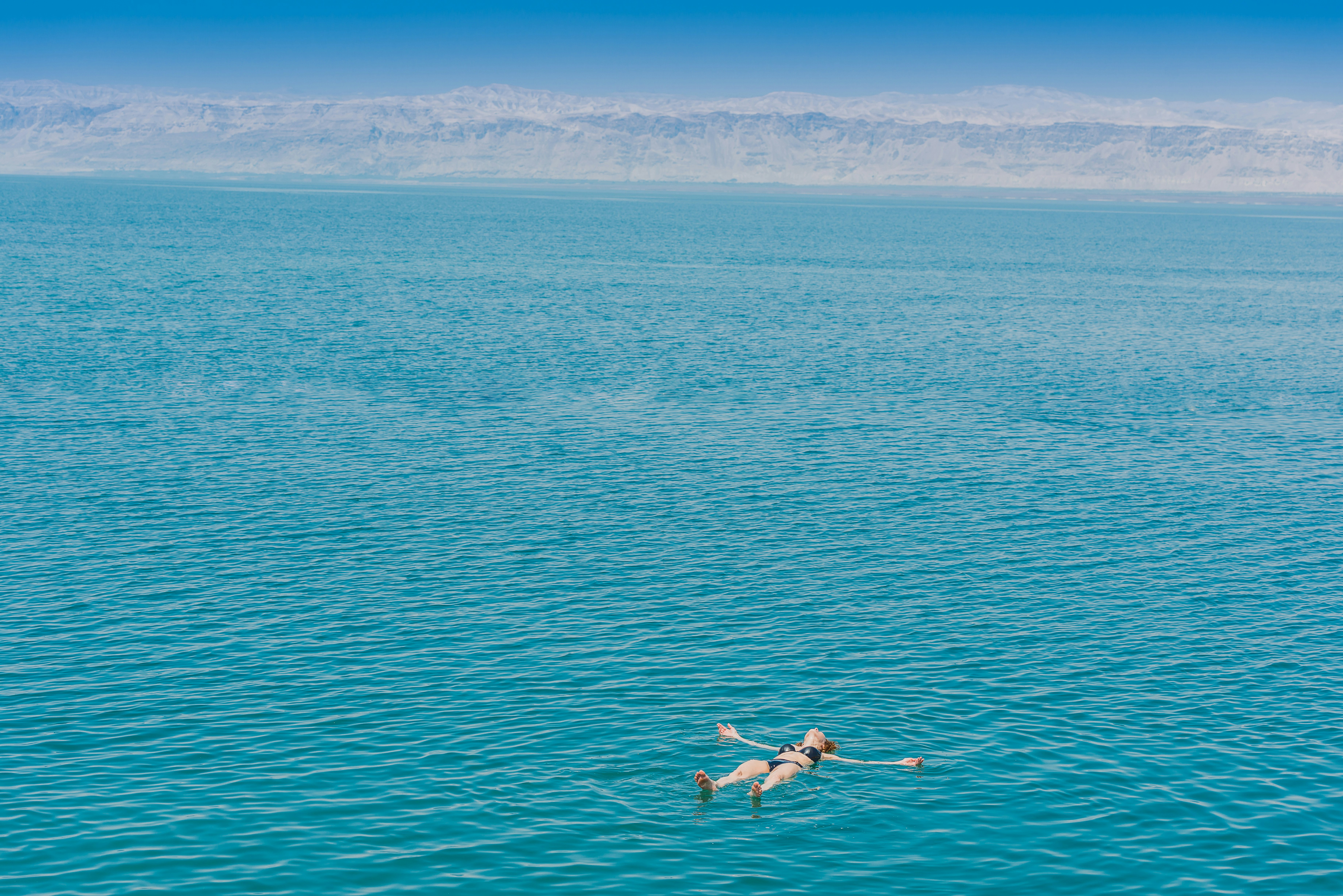 A vast blue lake backed by rocky mountains. A woman floats on the surface of the water with her arms out by her sides.