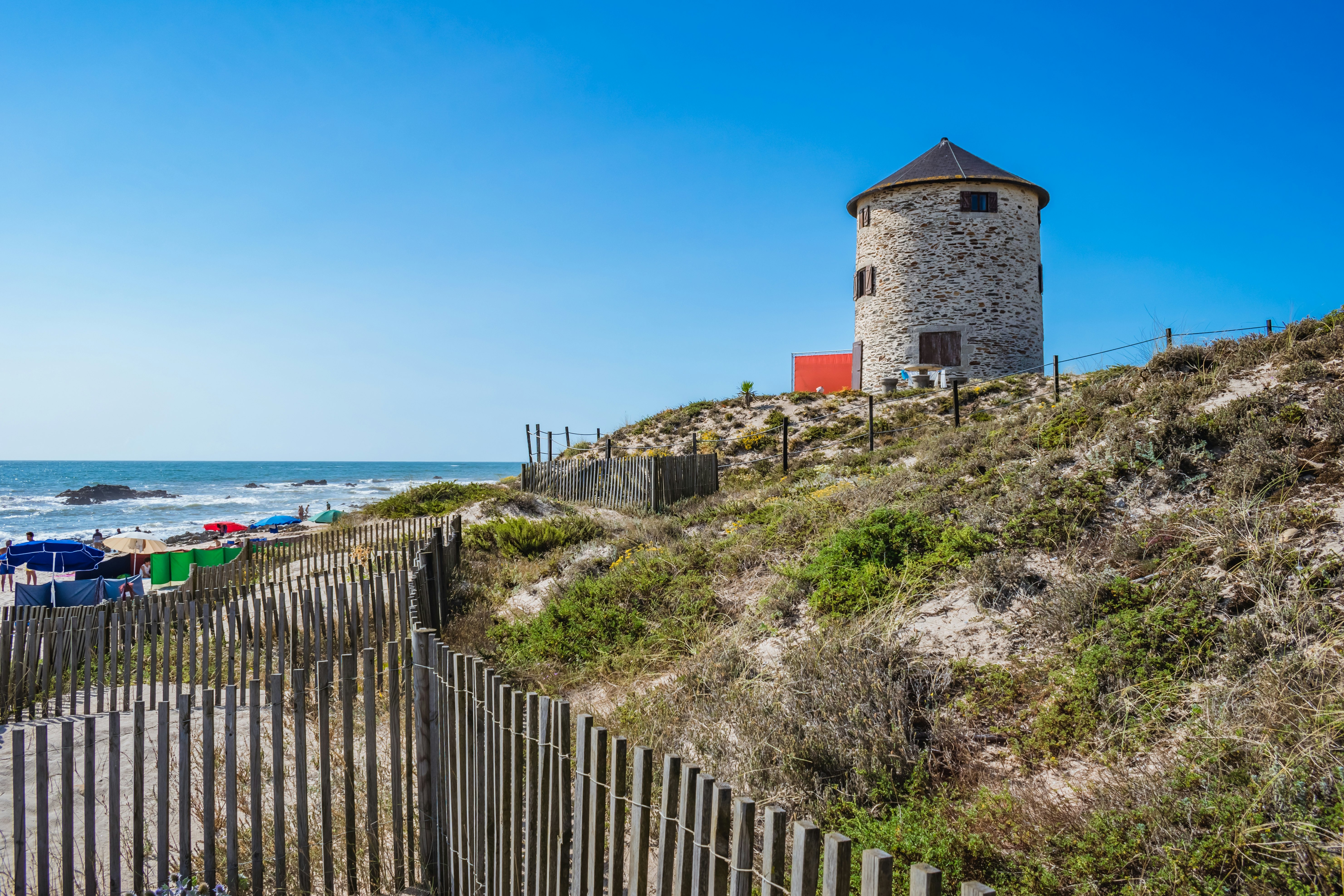 A stone windmill sits at the top of dunes by the ocean in Portugal. A wooden fence is in the foreground, and beach umbrellas are in the distance.